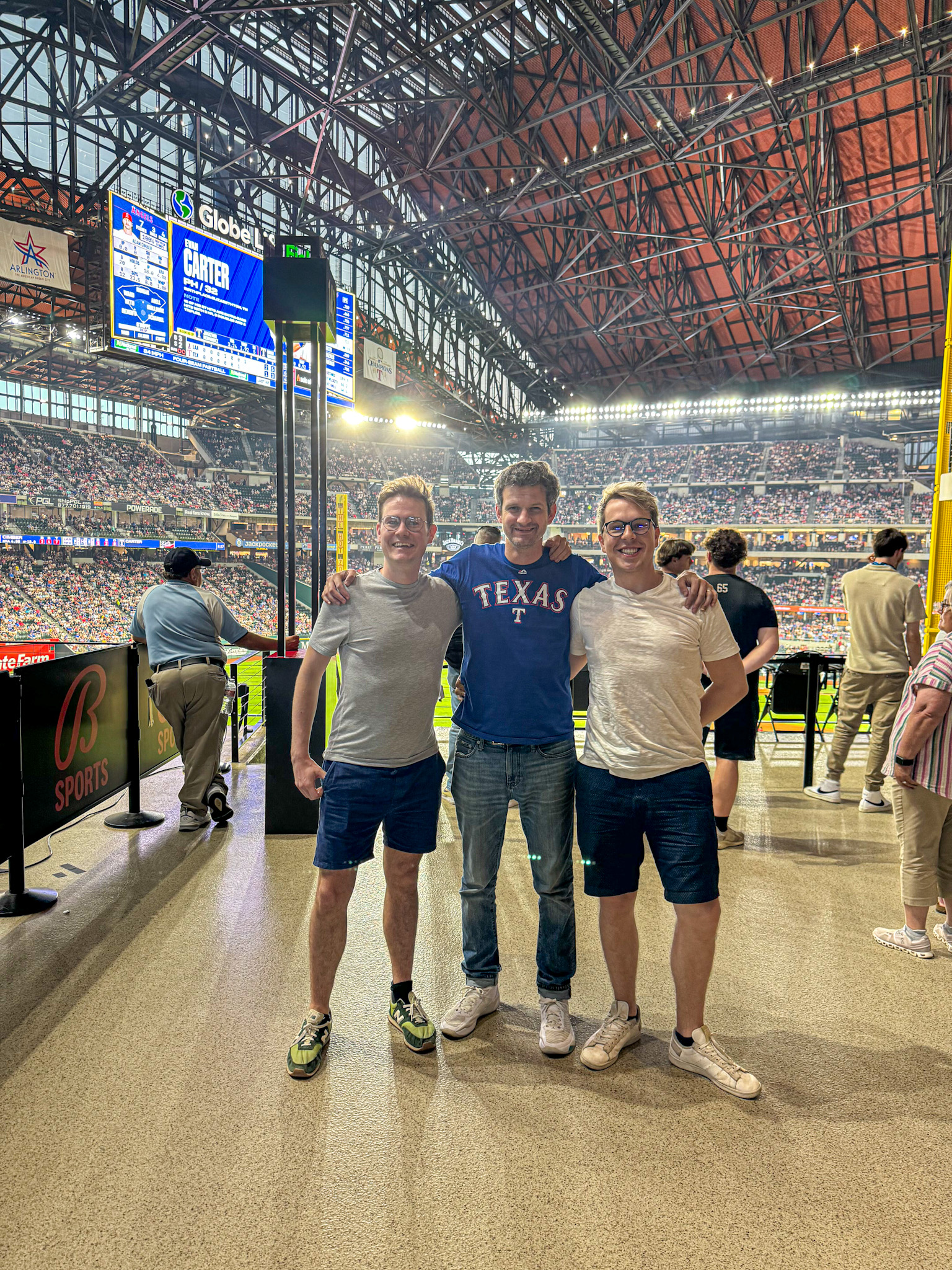 Three men smile and pose together inside a stadium during a baseball game at the Globe Life Field watching a Texas Rangers game in Arlington. 