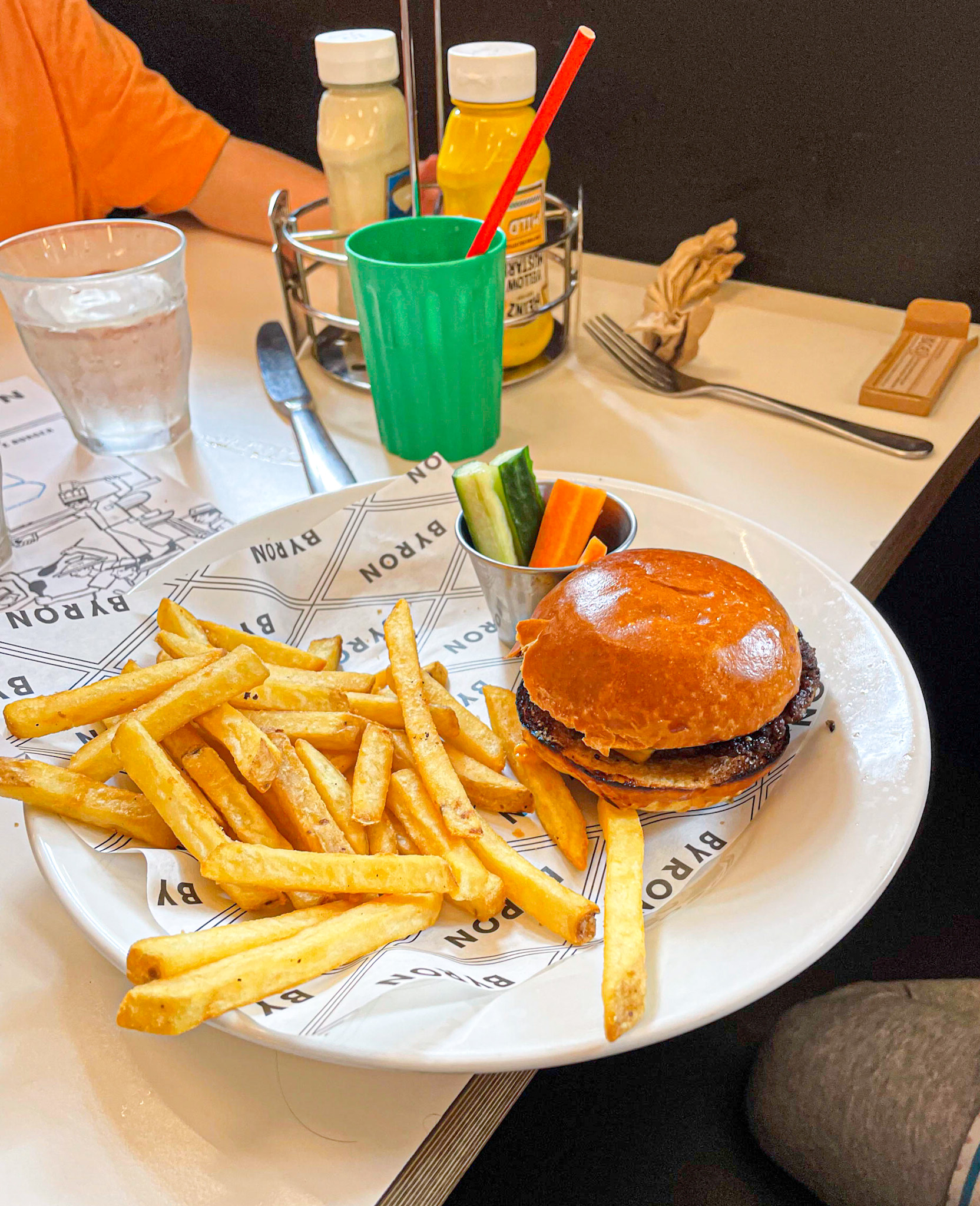A kid’s meal at Byron Burger featuring a cheeseburger, a generous serving of fries, and a cup of carrot and cucumber sticks. A green plastic cup with a red straw and condiments are on the table, with a partially colored activity sheet underneath the plate.