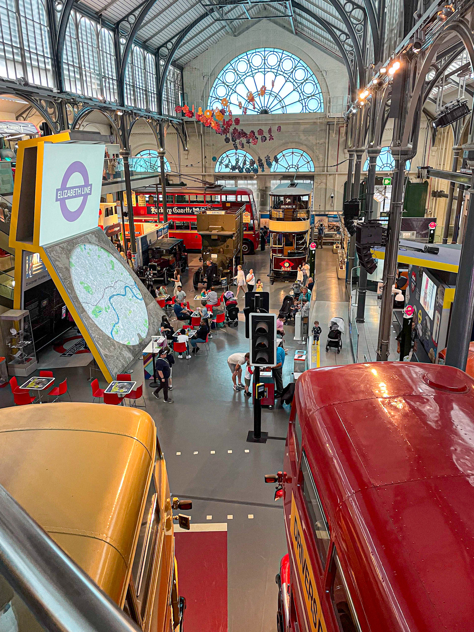 Interior view of the London Transport Museum featuring vintage red and yellow double-decker buses, interactive exhibits, and a large arched window. Visitors explore the space, with children playing near a life-size traffic light and families seated at activity tables below.