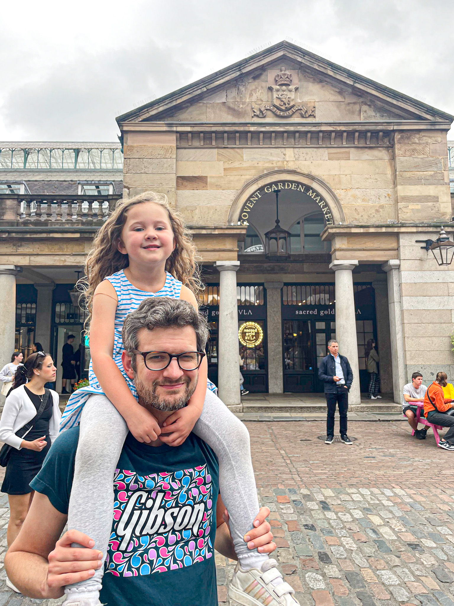 A man carries a smiling young girl on his shoulders in front of the Covent Garden Market entrance in London. The cobblestone square and historic building provide a charming backdrop as people walk and relax nearby.