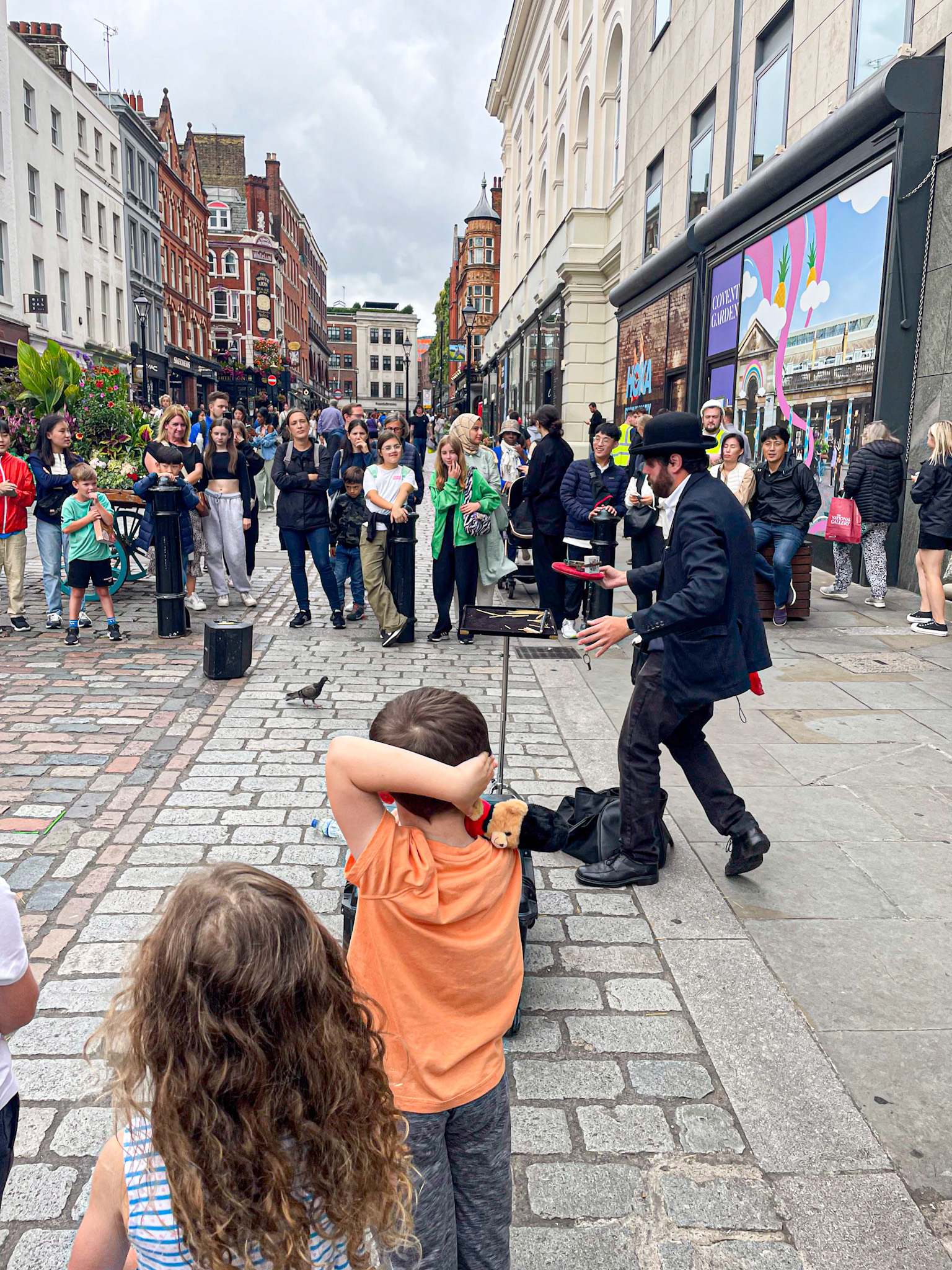 A lively street performer entertains a crowd on a cobblestone street in Covent Garden, London. Two children watch from the front row, one with hands on his head and a small toy tucked under his arm. The audience lines the street, surrounded by historic buildings and colorful storefronts.