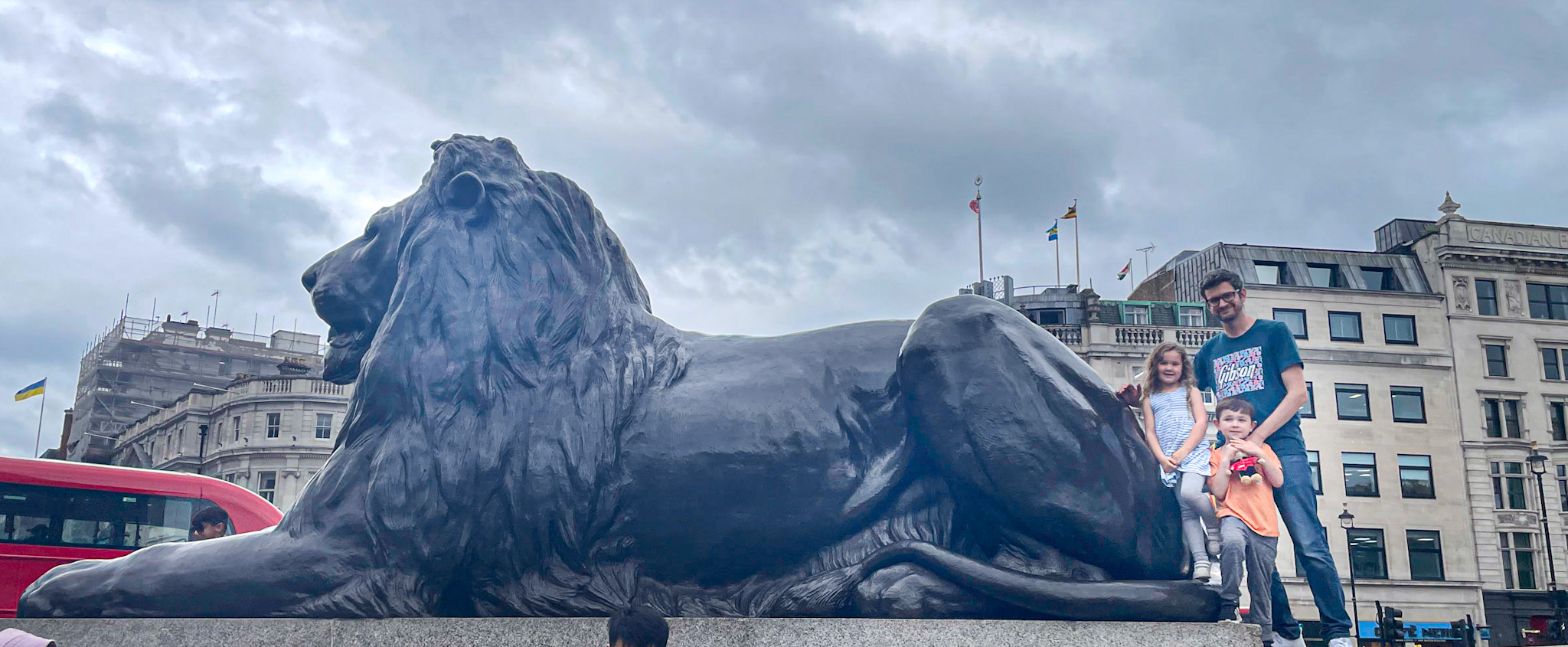 A father and two young children stand beside one of the massive lion statues in Trafalgar Square, London. The iconic black lion dominates the foreground while London buildings, flags, and a red double-decker bus appear in the background under cloudy skies.