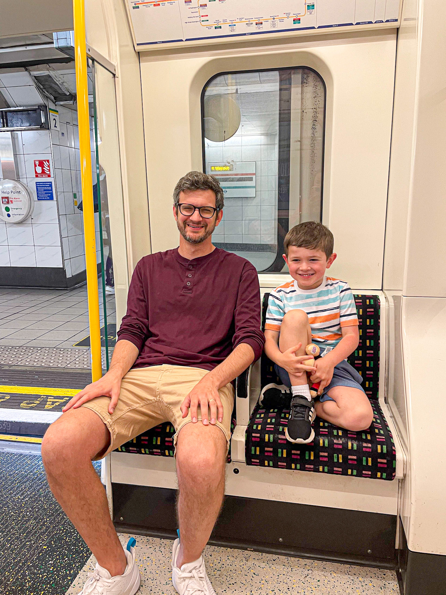 A smiling man and young boy sit side by side on a London Underground train, enjoying the ride. The man wears a maroon shirt and khaki shorts, while the boy is in a striped t-shirt and shorts, holding a toy. The train is stopped at a station, visible through the open door behind them.