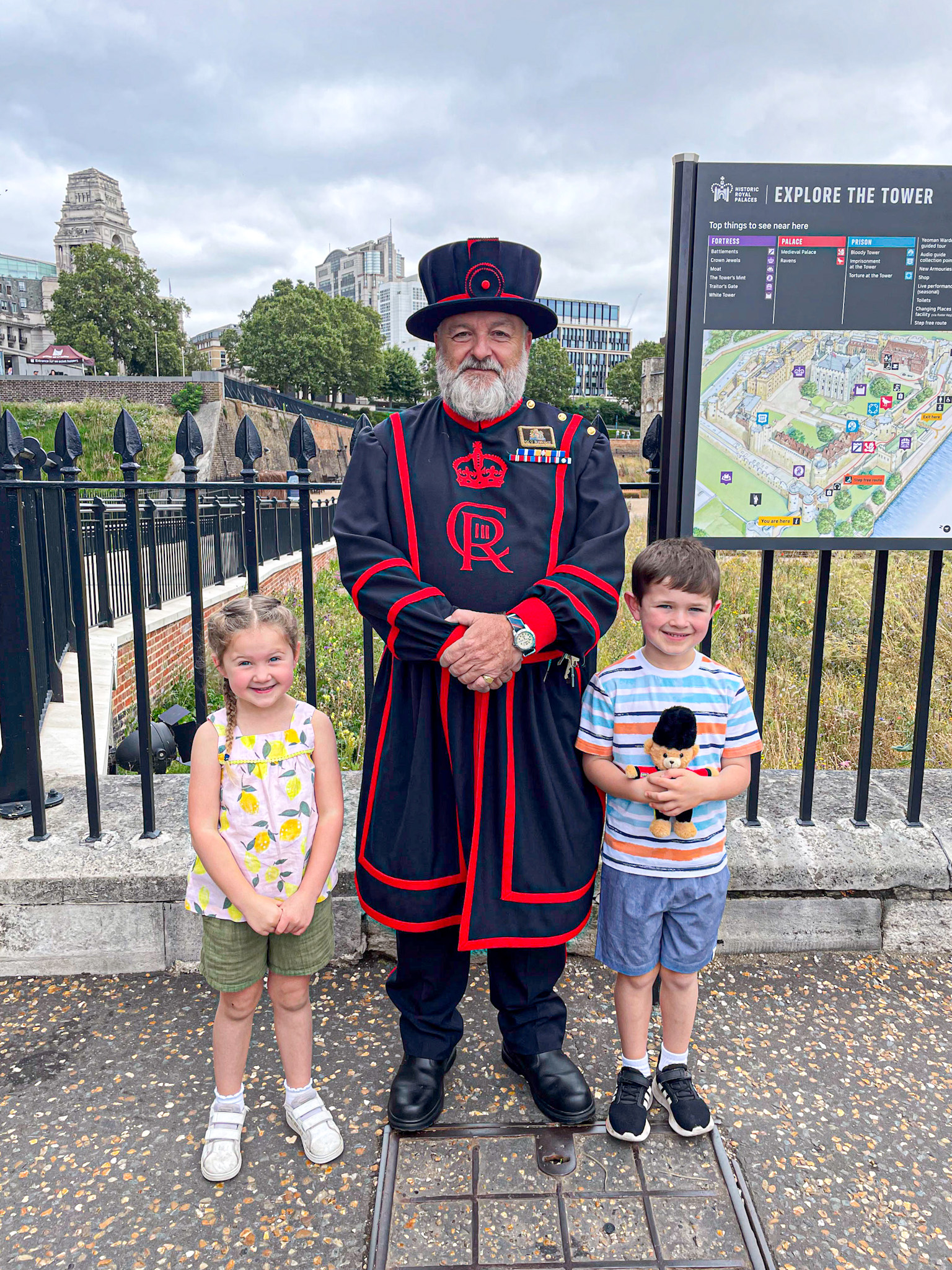 Two smiling children pose with a Beefeater guard at the Tower of London. The guard wears a traditional dark uniform with red trim and a tall hat, while the boy holds a small toy guardsman. A sign reading “Explore the Tower” and part of the London skyline are visible in the background.
