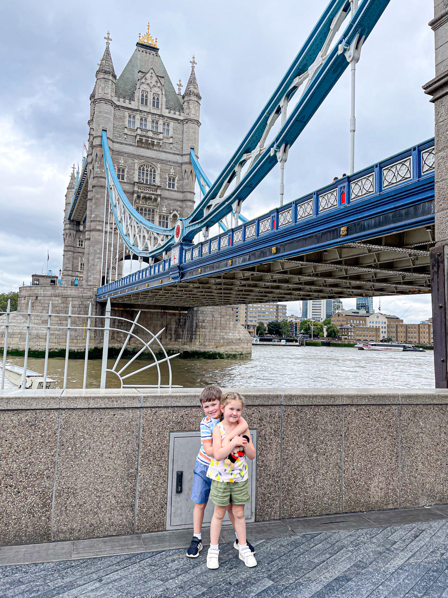 Two young children hug and smile in front of Tower Bridge in London, with the iconic blue and white suspension bridge and one of its towers rising behind them. The River Thames and city buildings are visible in the background under a cloudy sky.