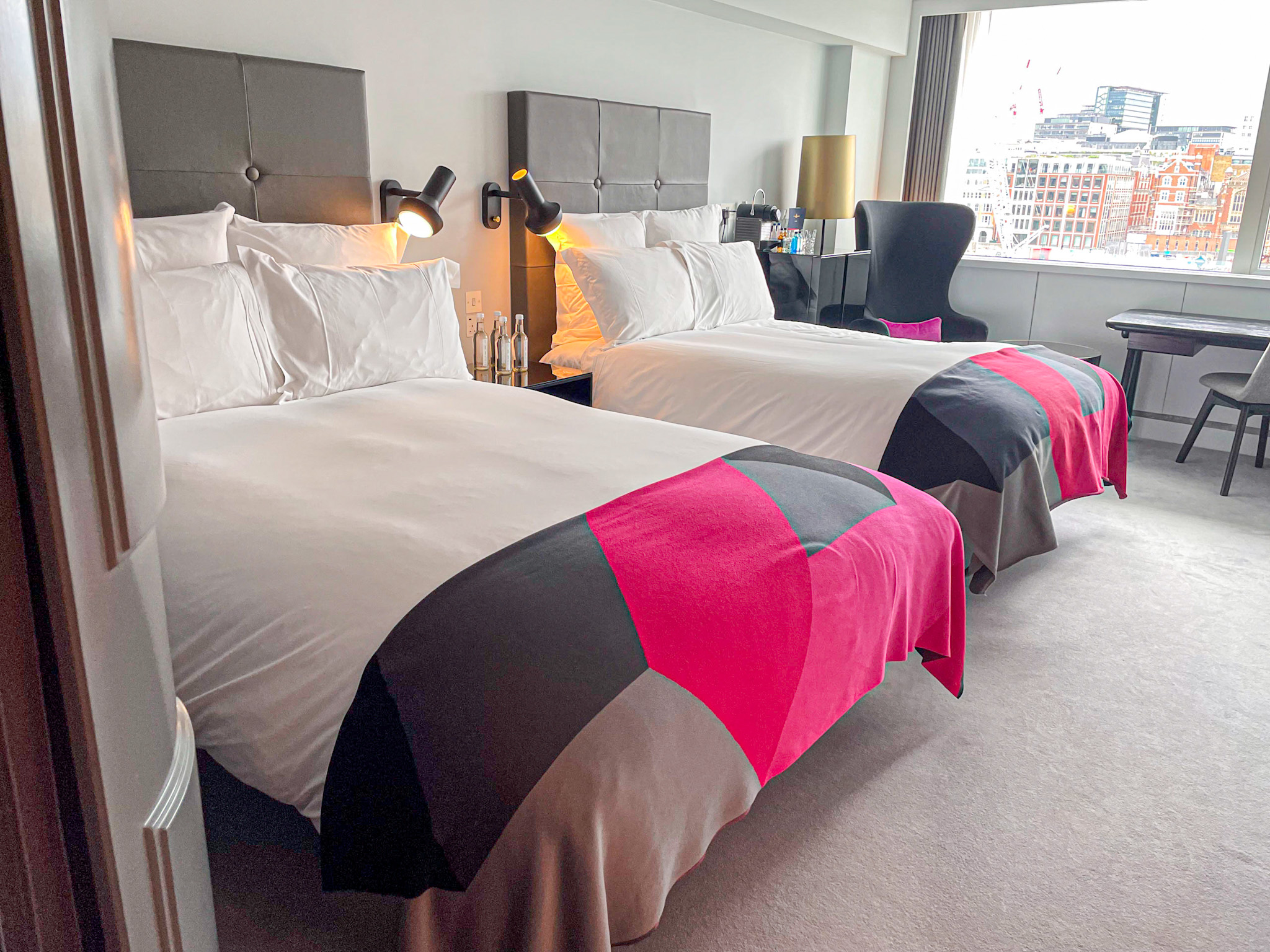 Two plush double beds with crisp white linens and bold pink, black, and gray throws in a modern hotel room at Sea Containers London, featuring large windows with a city view, a work desk, and cozy seating.