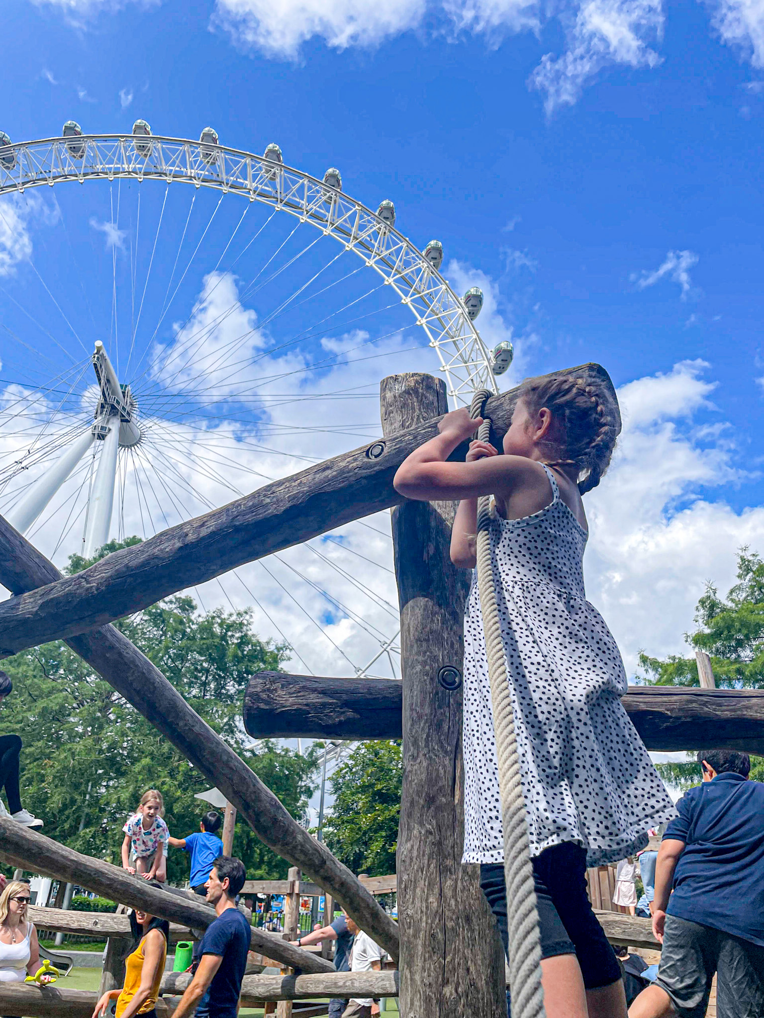 A young girl climbs a rope structure at a wooden playground with the London Eye towering in the background against a bright blue sky. Other kids and parents are scattered around enjoying the sunny day.