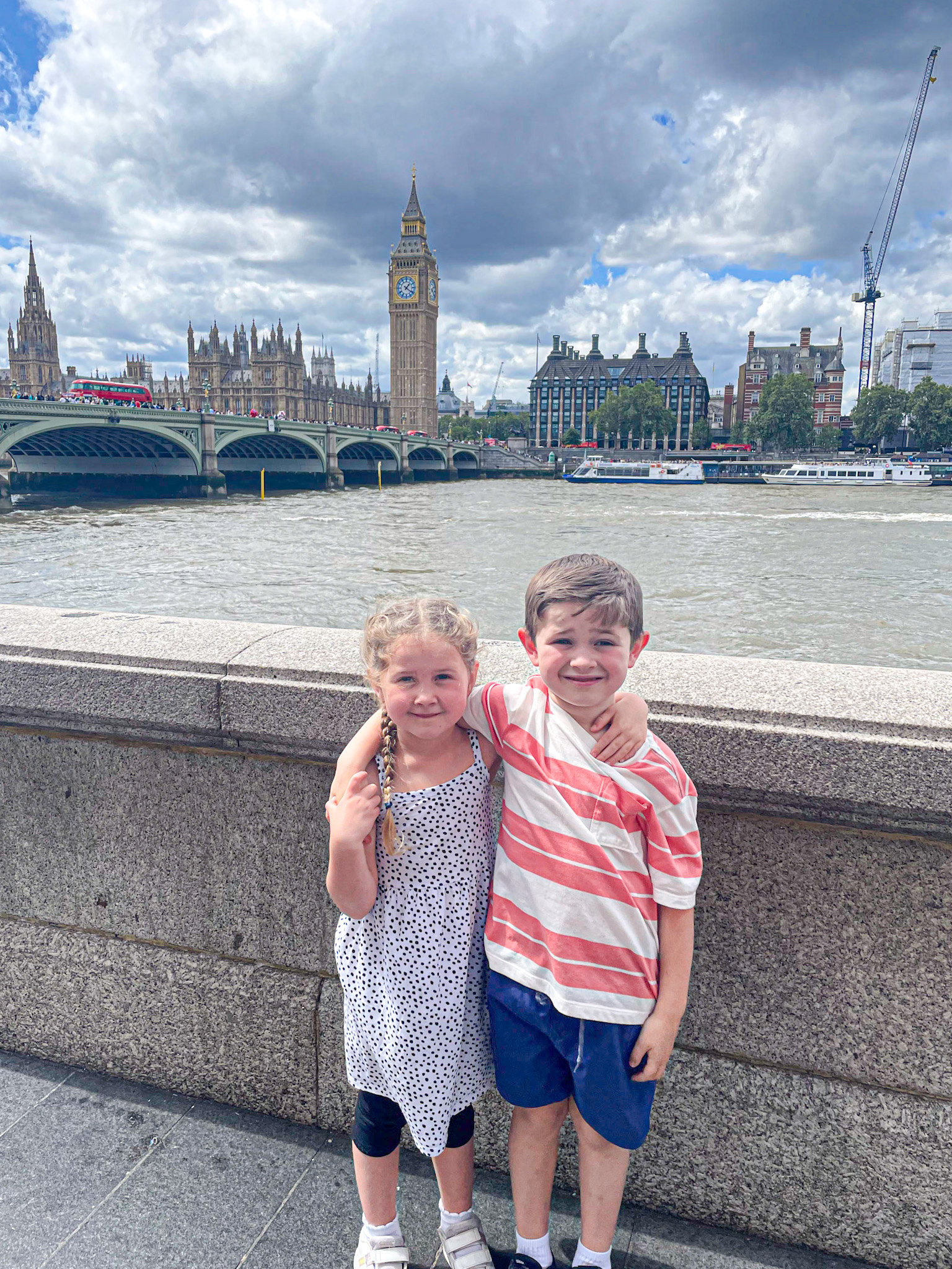 Two smiling children pose arm-in-arm in front of the River Thames with Big Ben and the Houses of Parliament in the background. The girl wears a white polka dot dress with black leggings and the boy wears a red and white striped shirt with blue shorts. A red double-decker bus crosses Westminster Bridge behind them under a dramatic, cloud-filled sky.