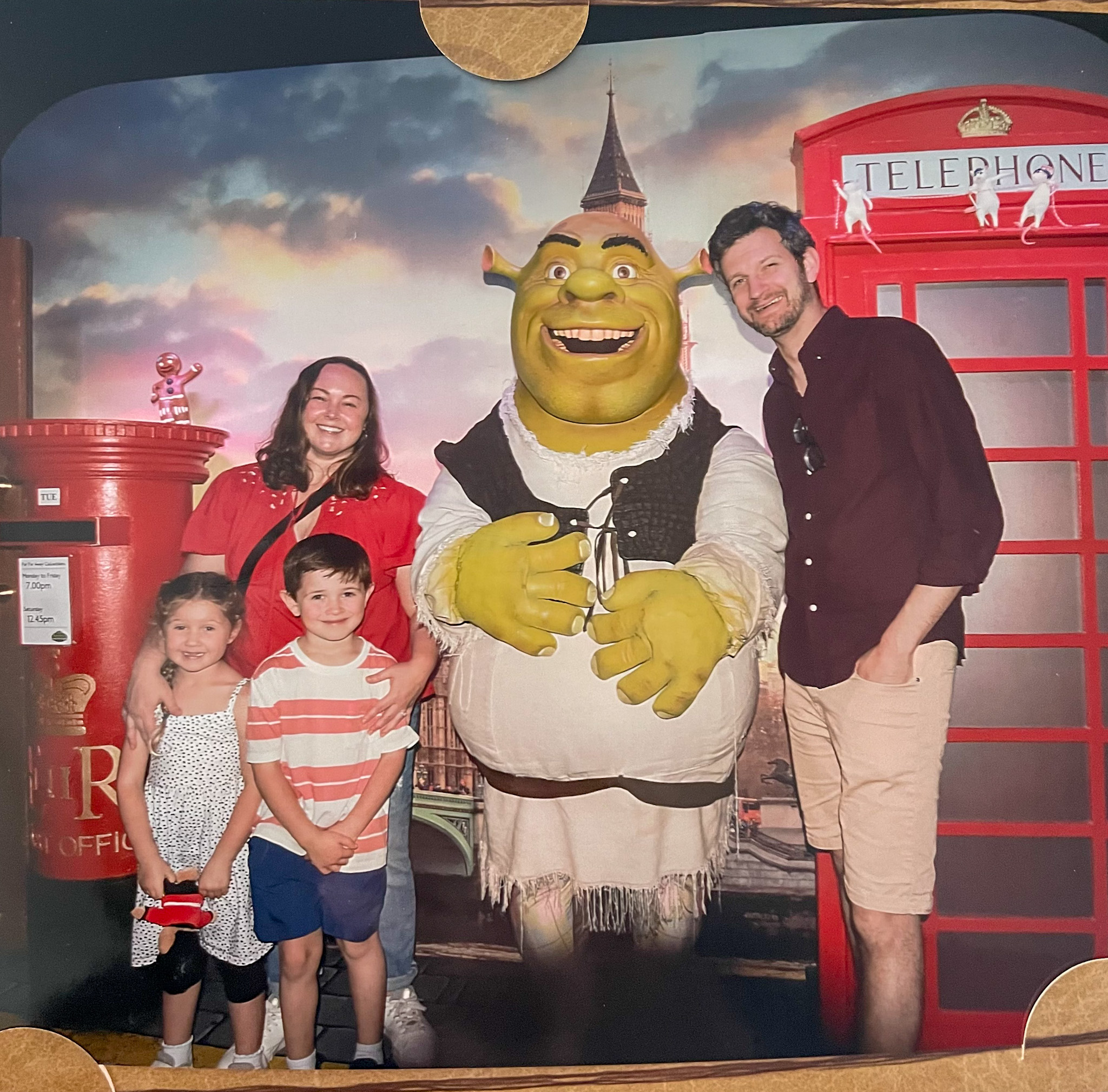 A smiling family of four poses with a life-sized Shrek character at Shrek’s Adventure London. Behind them is a colorful backdrop featuring a red British telephone booth, a royal mail postbox, and Big Ben. The kids look excited, and everyone is dressed for a fun day out.