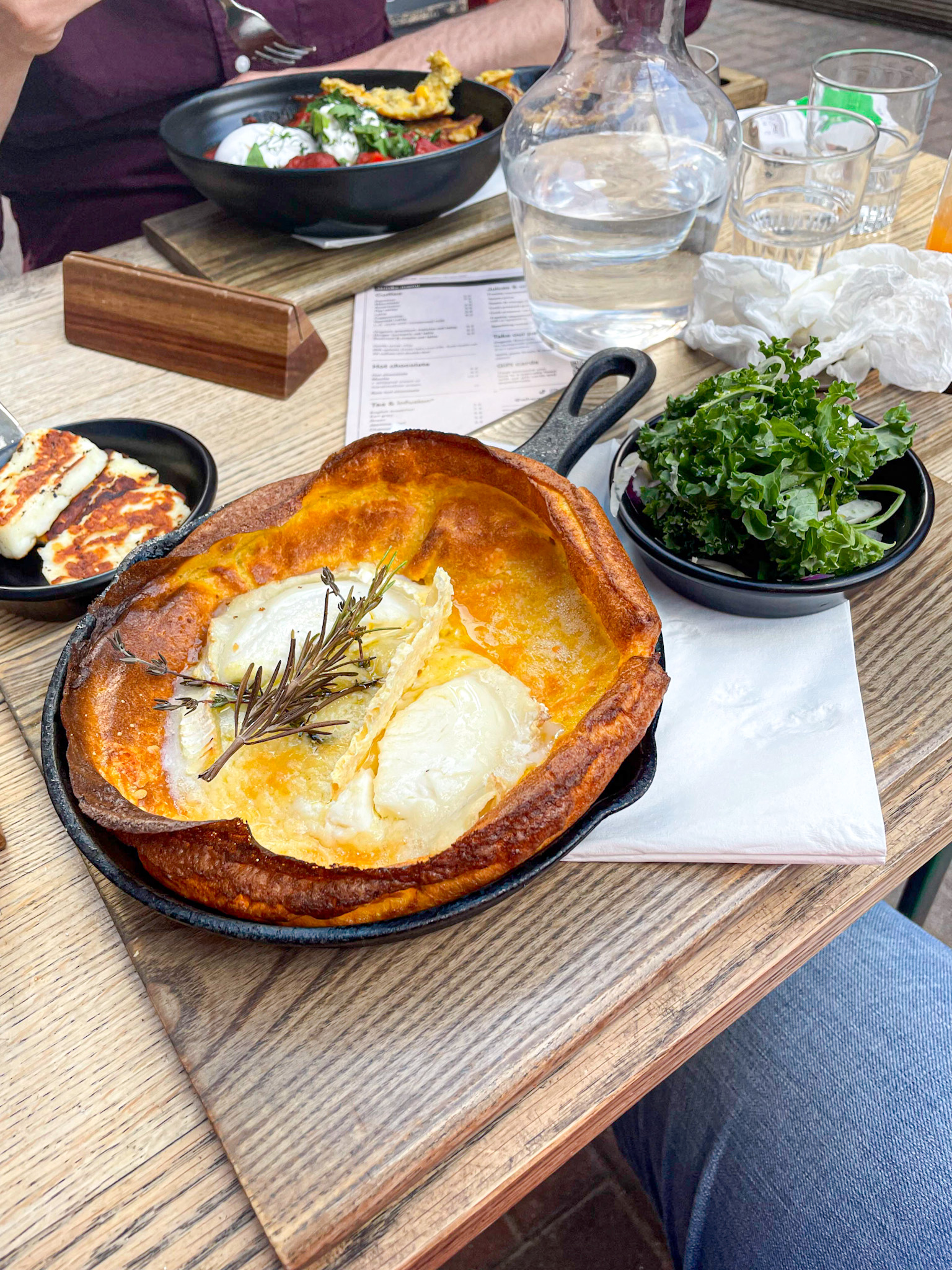 A skillet-baked Yorkshire pudding filled with melted cheese and topped with a sprig of rosemary sits on a wooden table next to a small bowl of fresh kale salad. In the background, other brunch dishes and drinks are visible.
