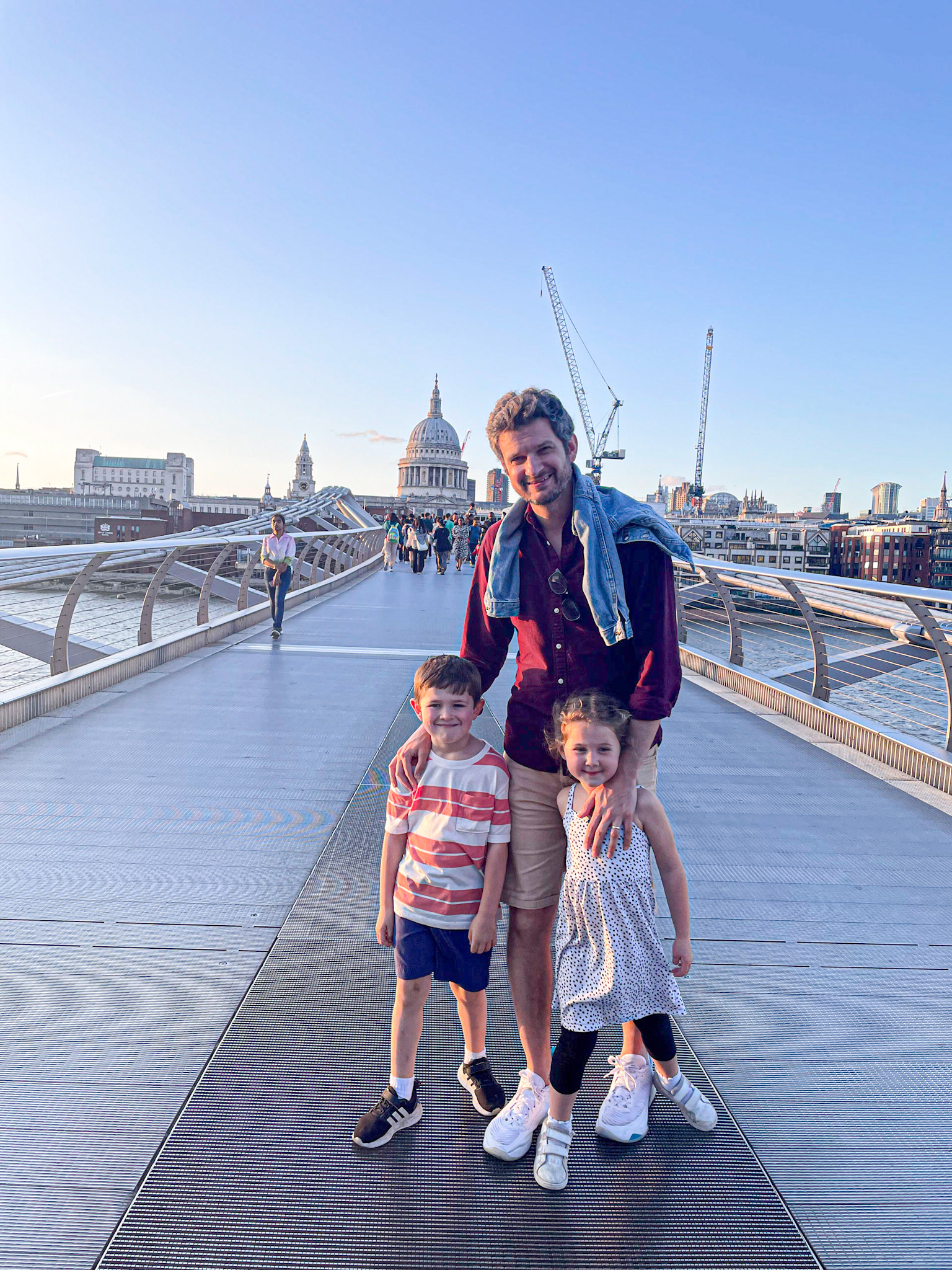 A man and two smiling children stand on the Millennium Bridge in London at sunset, with St. Paul’s Cathedral clearly visible in the background. The bridge is lightly crowded with pedestrians, and cranes rise above the city skyline under a clear blue sky.