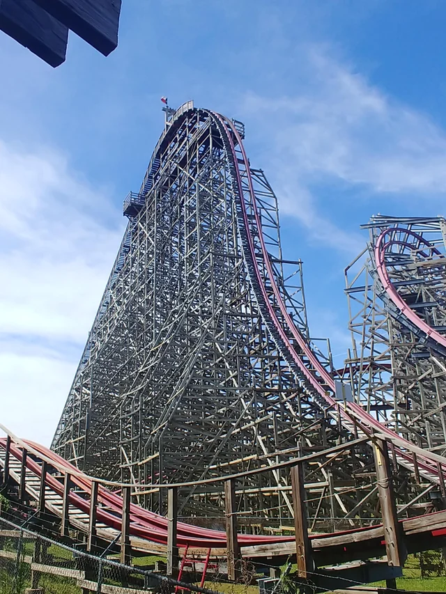 A large wooden roller coaster at Six Flags in Arlington with red tracks rises sharply against a blue sky with scattered clouds.