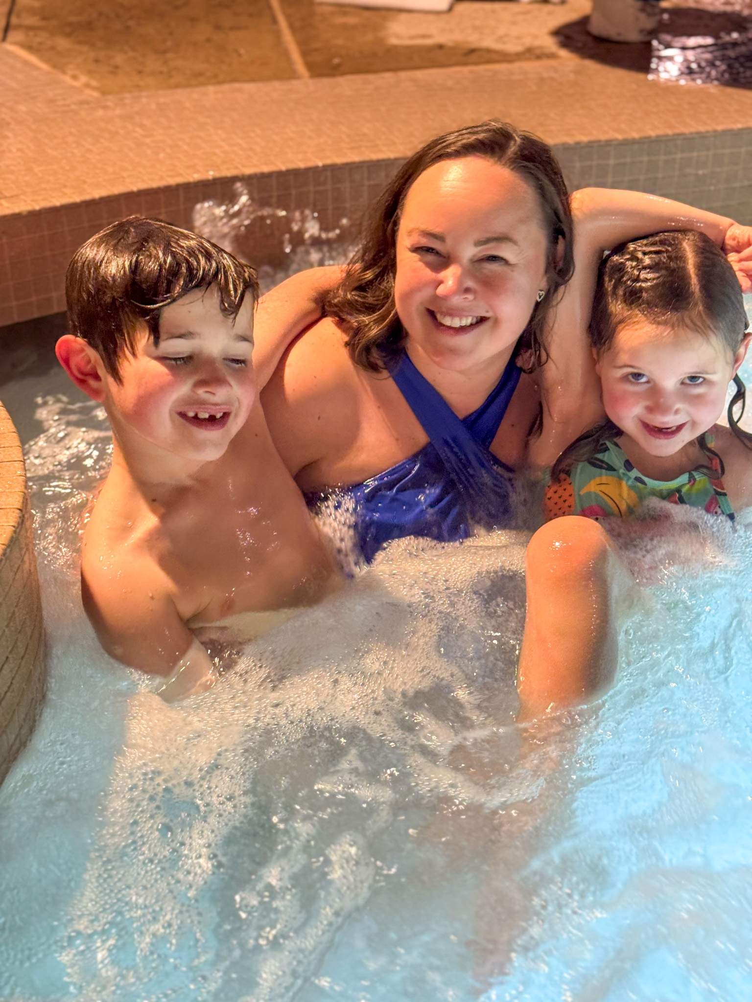 A woman and two young children smile while sitting together in a bubbling hot tub.