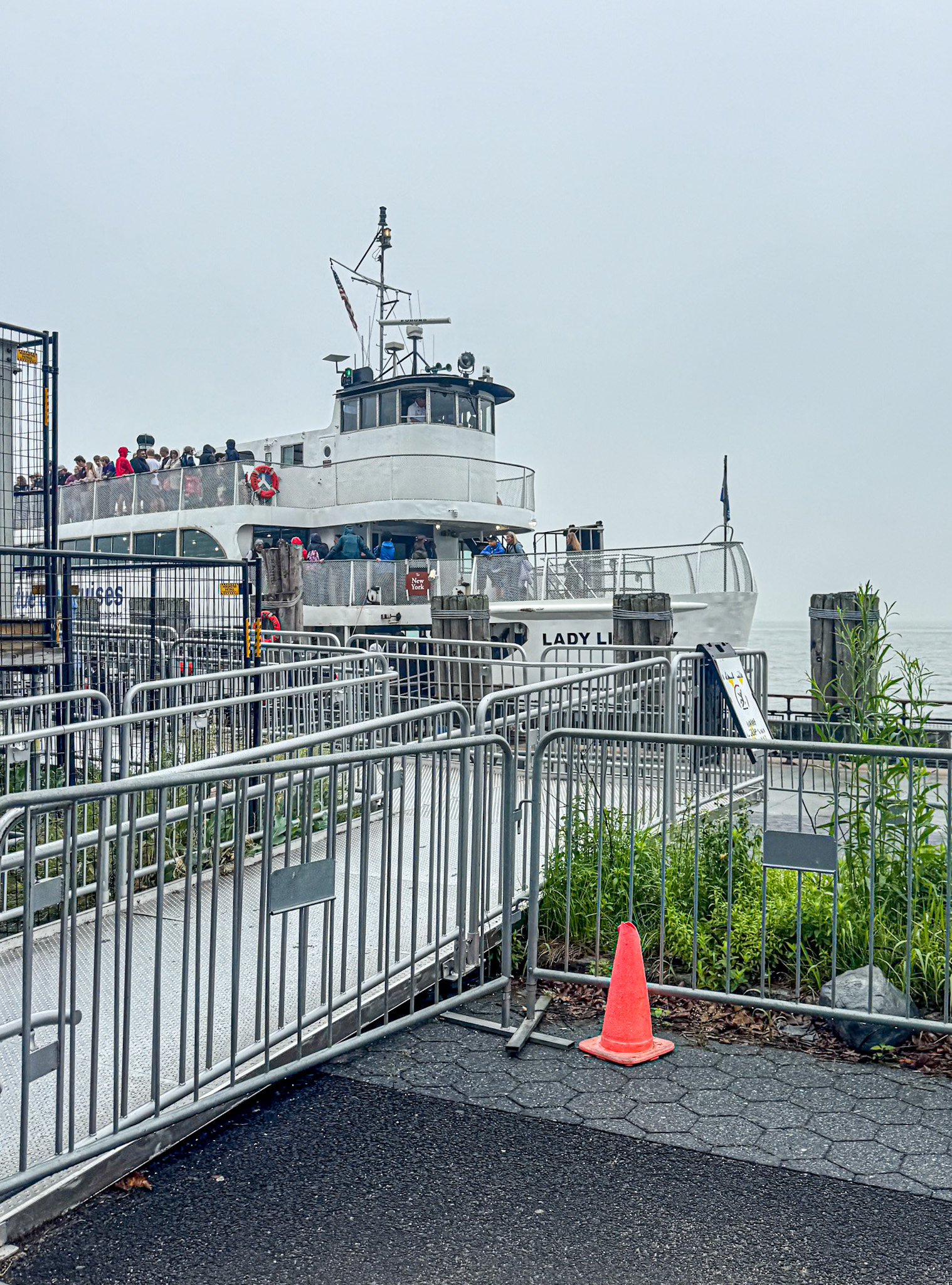 A ferry named Lady Liberty docked, with passengers on board and metal railings in the foreground at Battery Park in Manhattan.