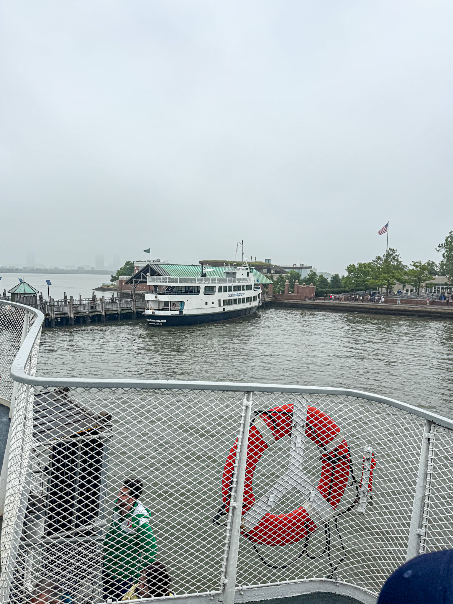 View from a statue of liberty ferry with a life preserver, looking toward a docked boat and buildings on a cloudy day.