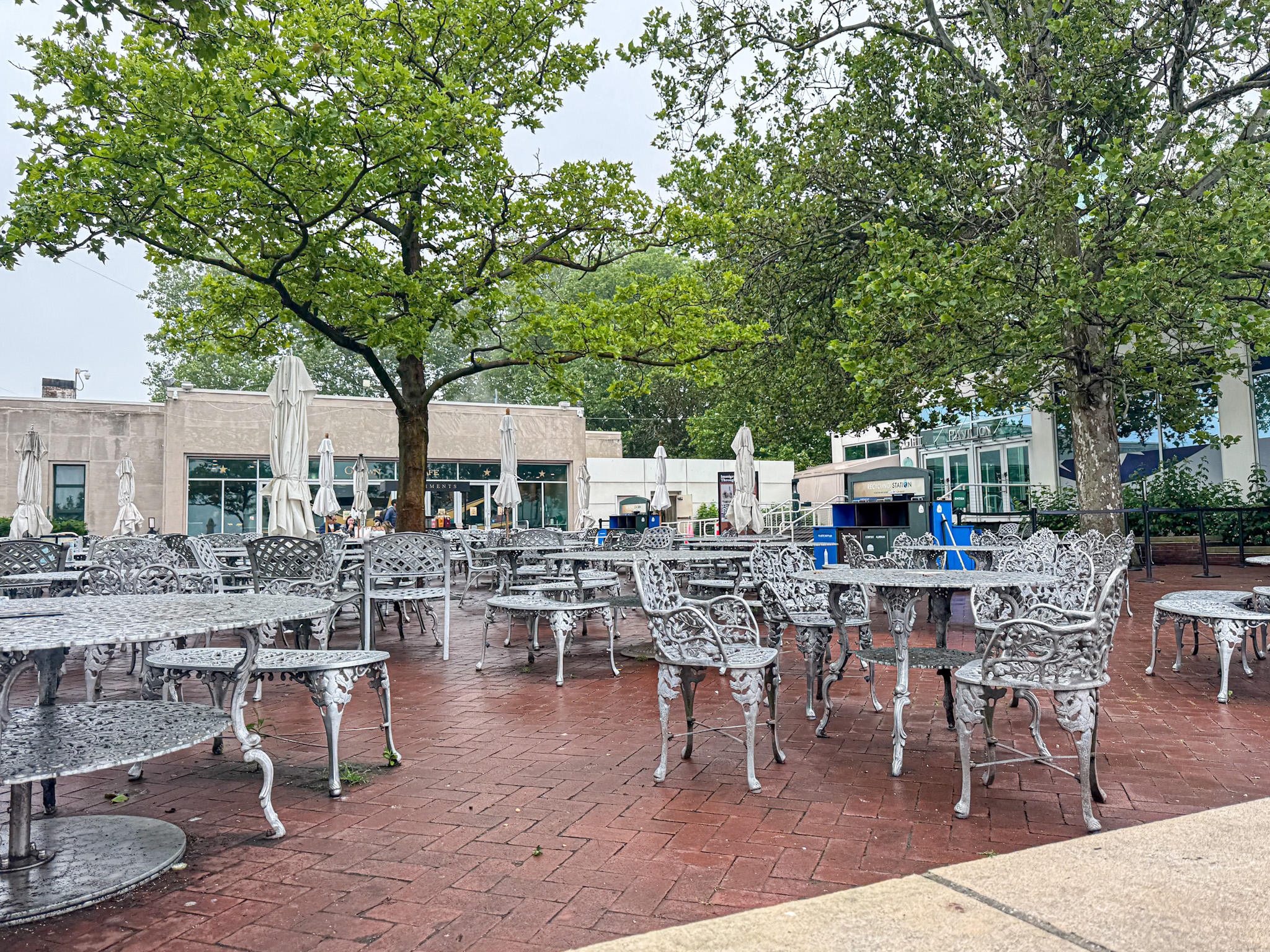 Outdoor patio with ornate metal tables and chairs under leafy trees on a brick surface, no people present.