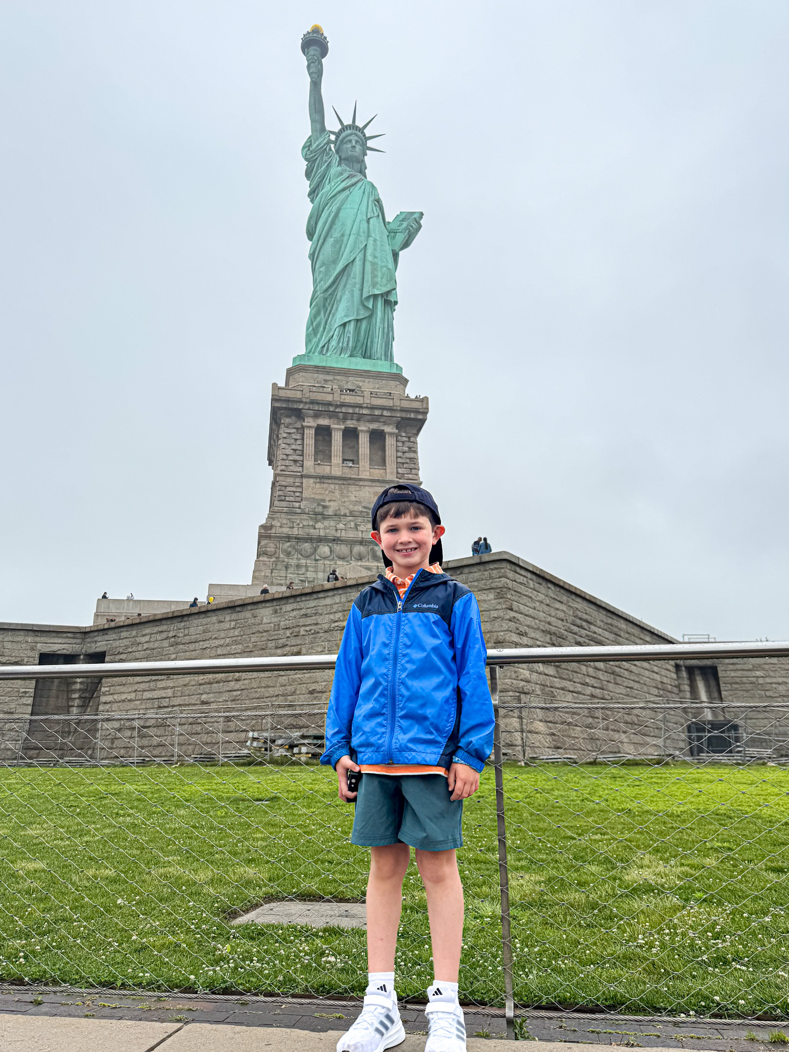 A smiling boy stands in front of the Statue of Liberty on a cloudy day.