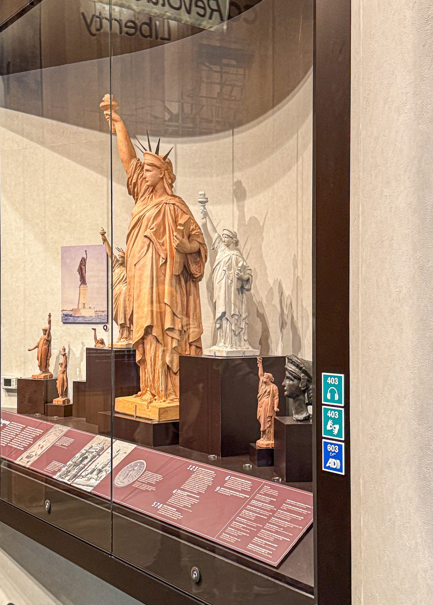 Miniature Statue of Liberty models are displayed behind glass in a museum exhibit with informational placards.