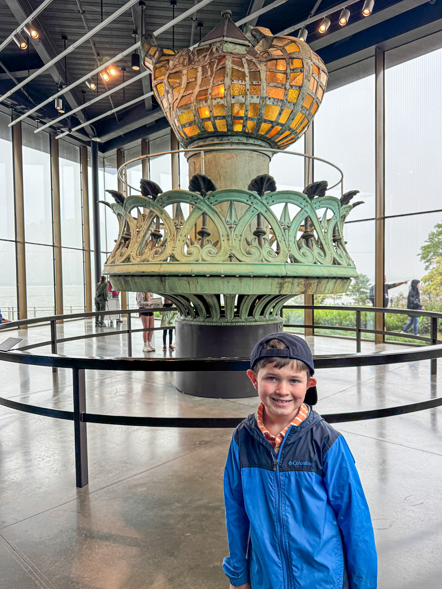 A young boy smiles in front of the original Statue of Liberty torch displayed indoors behind glass barriers.