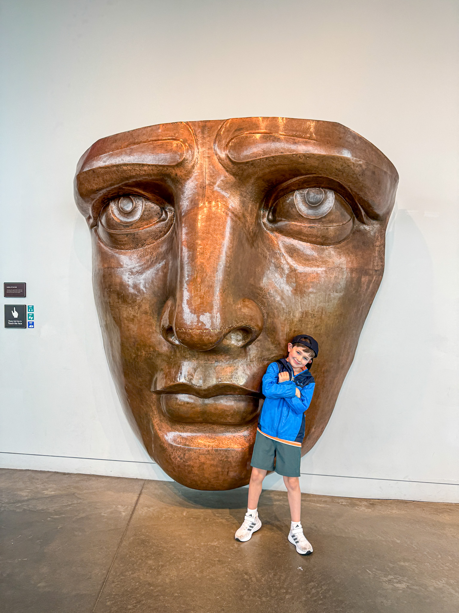 A child poses in front of a large bronze sculpture of a Lady Liberty's face mounted on a wall. 