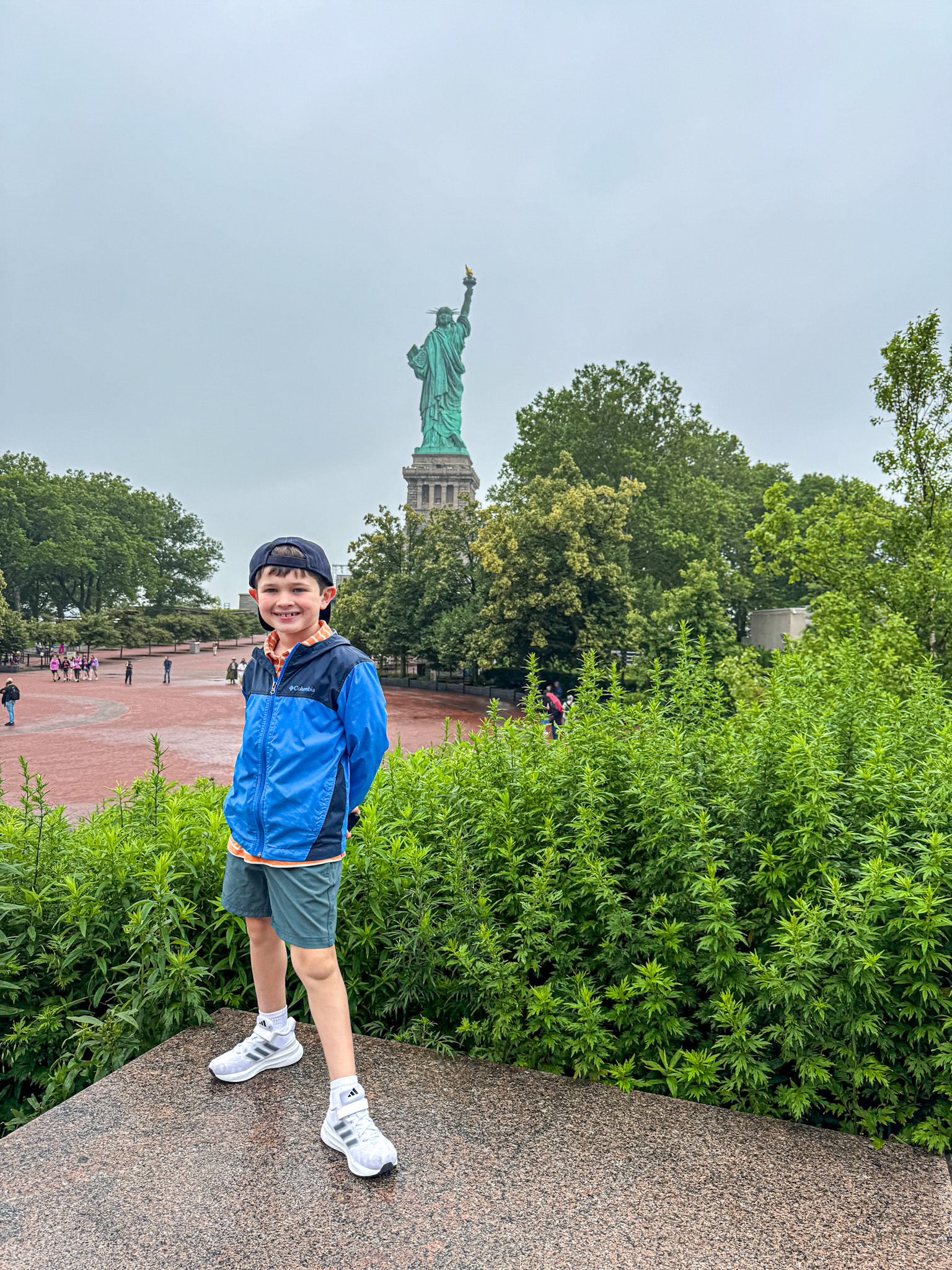 Smiling boy poses in front of the Statue of Liberty on a cloudy day, surrounded by greenery.
