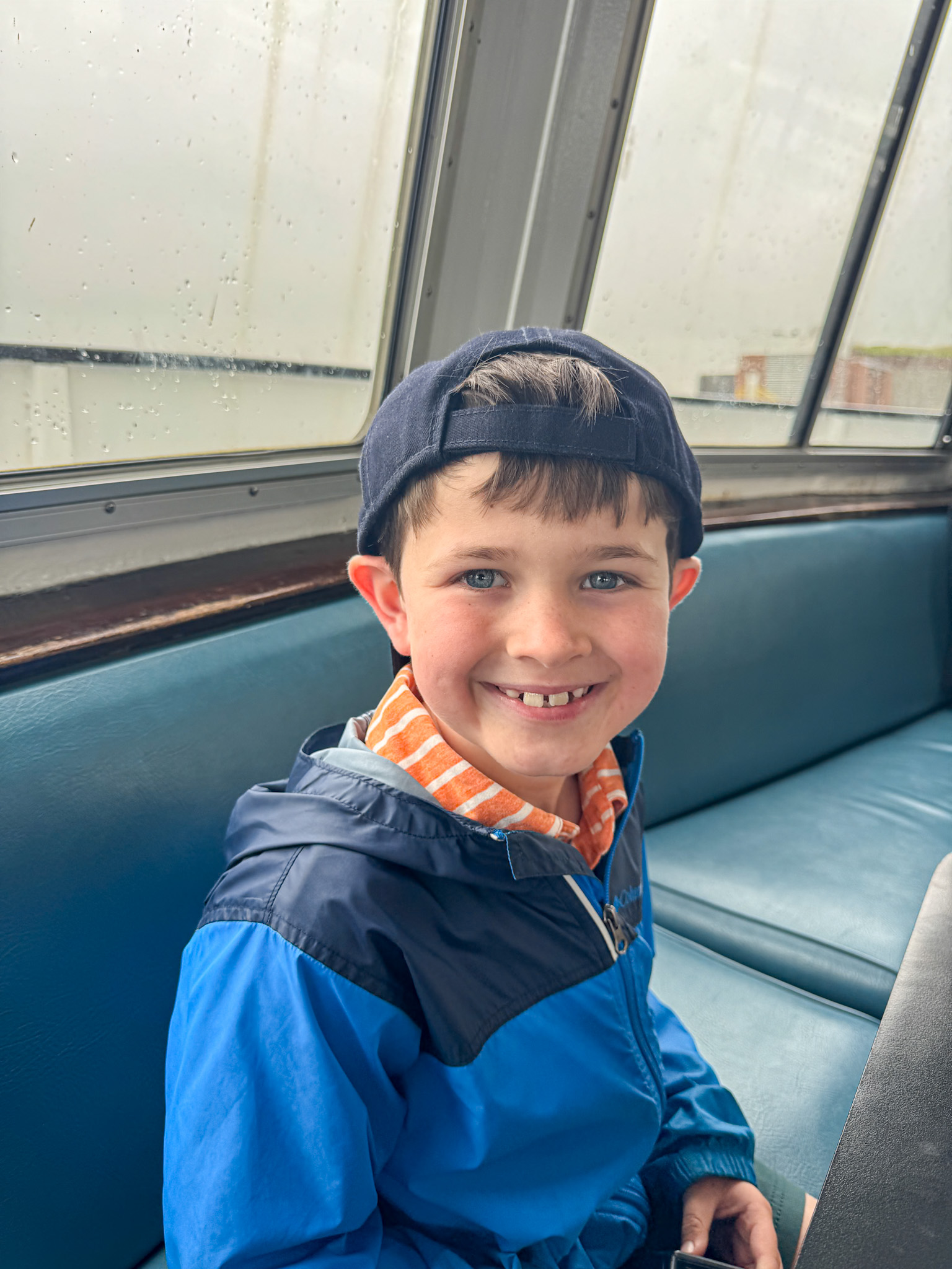 Smiling boy in a blue jacket and cap sitting on a blue bench inside a boat to Liberty Island. 