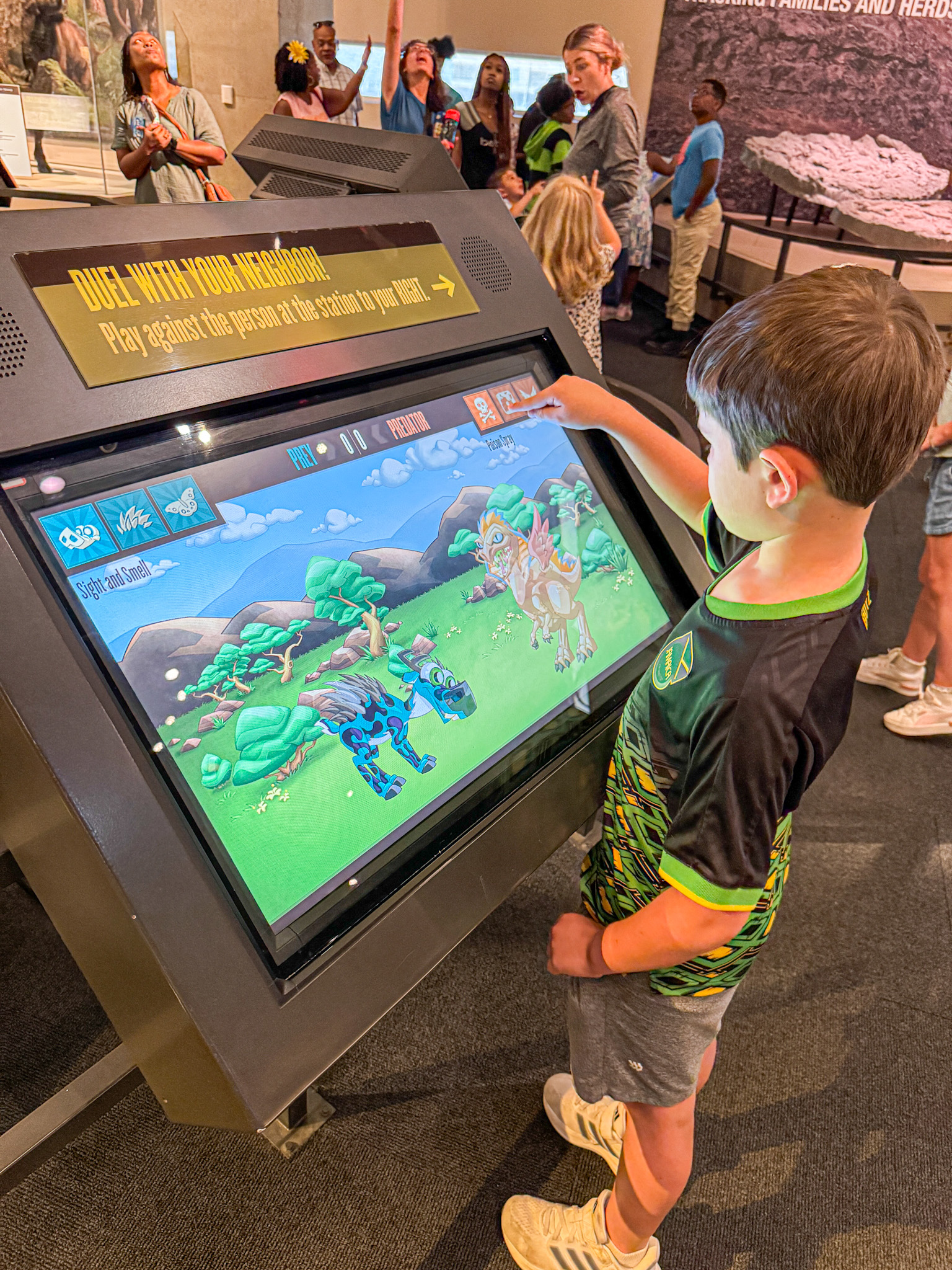 A boy uses a large touchscreen game at a museum while other visitors look on in the background.
