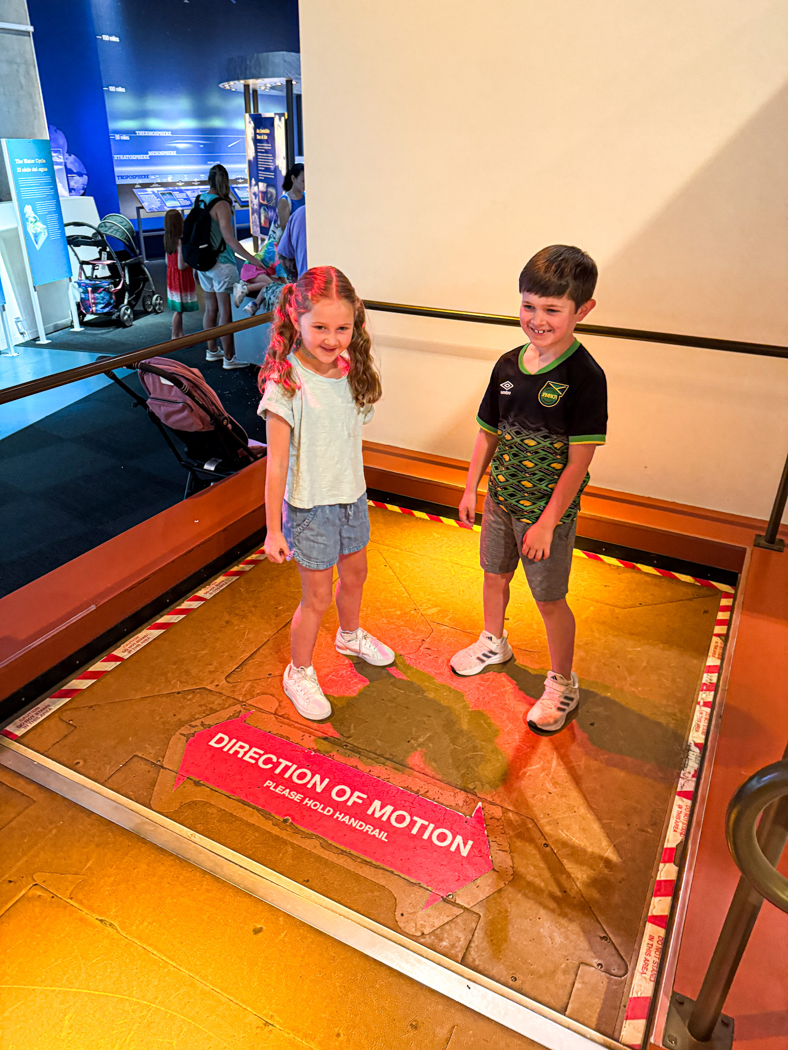 Two young children smile while standing on a earthquake simulator at the Rees-Jones Foundation Dynamic Earth Hall.