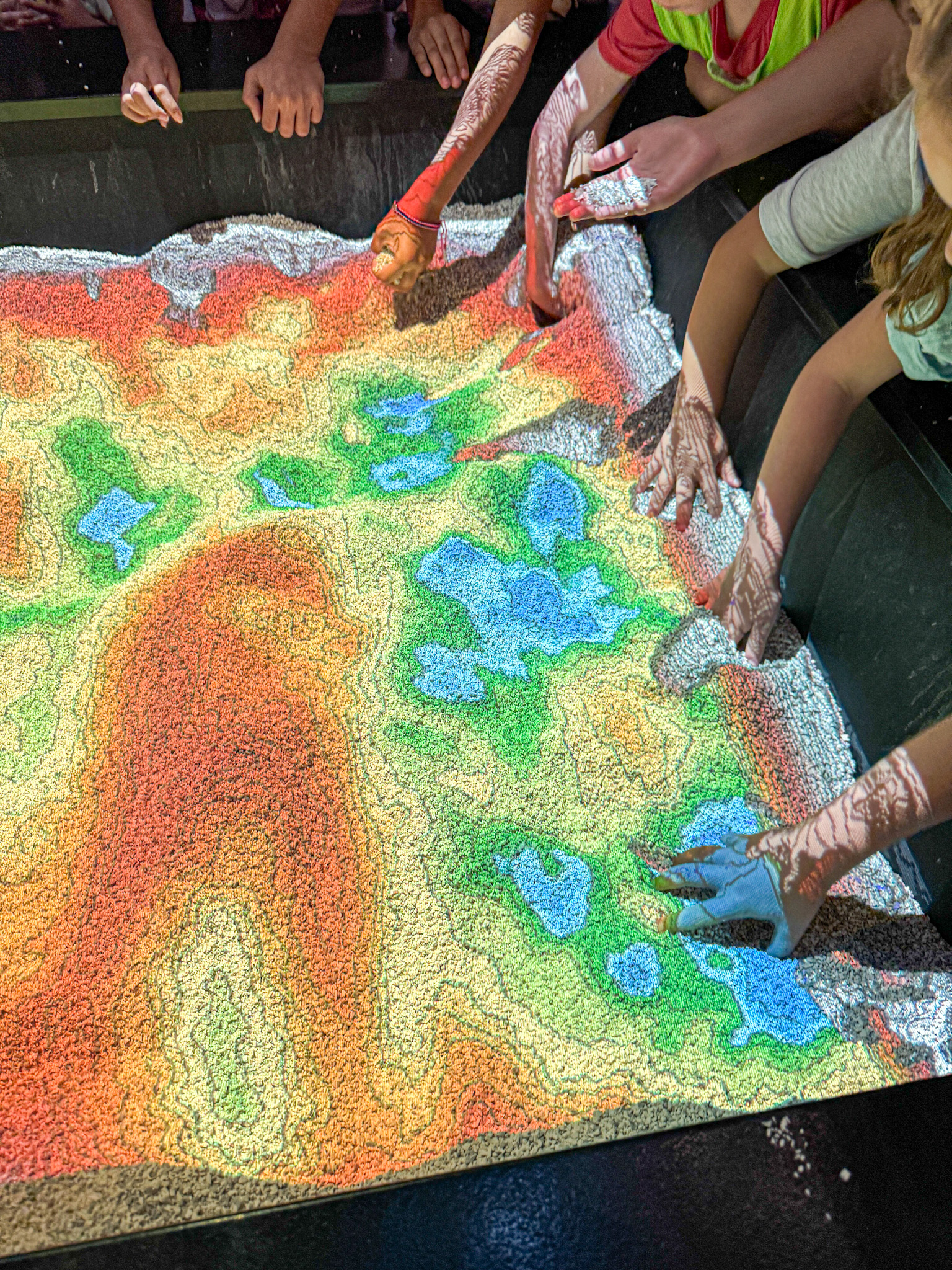 Children use their hands to shape a colorful interactive sandbox with projected topographic map patterns.
