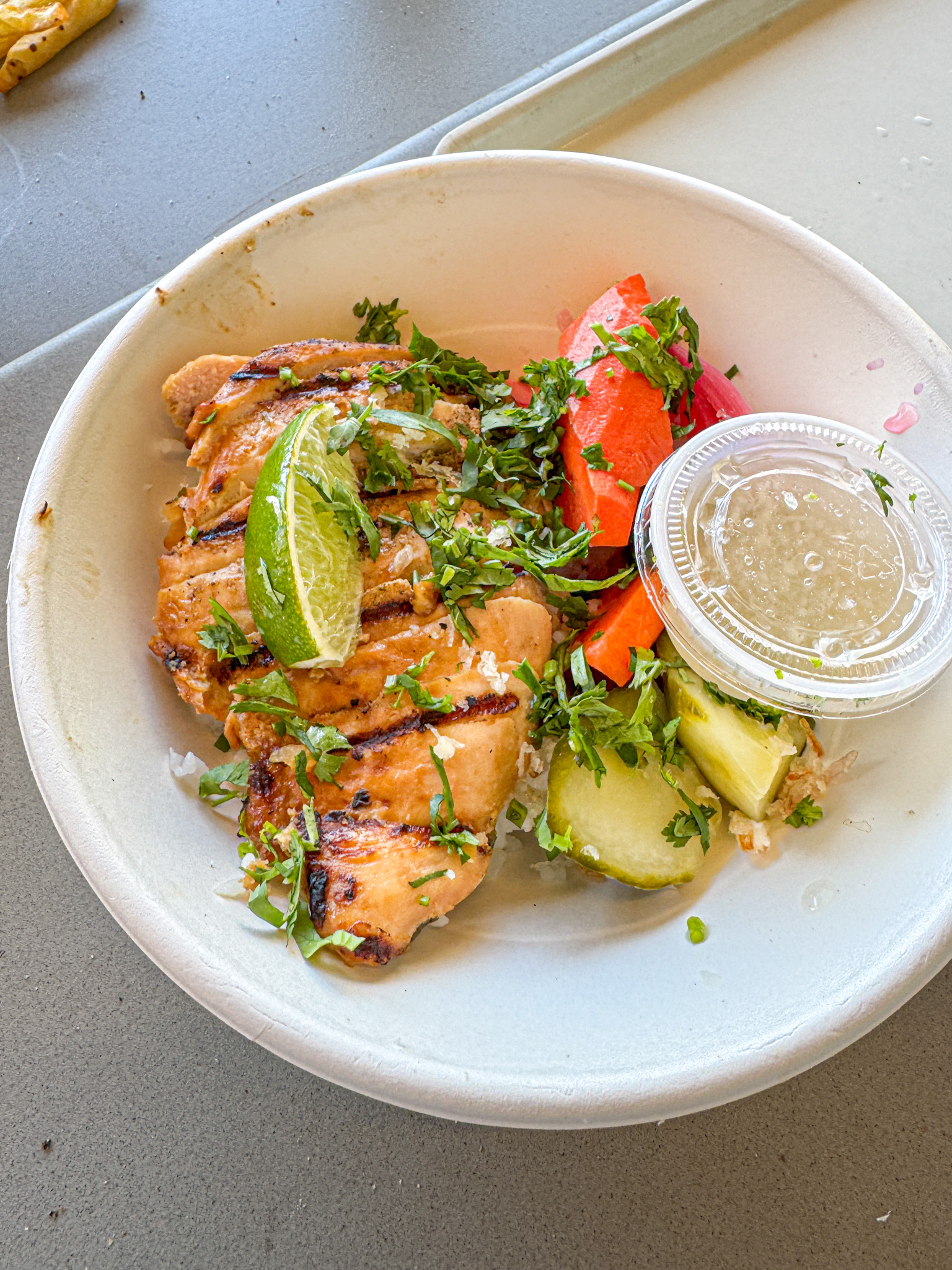 Grilled chicken with lime, fresh herbs, pickled veggies, and dressing in a white bowl on a table.