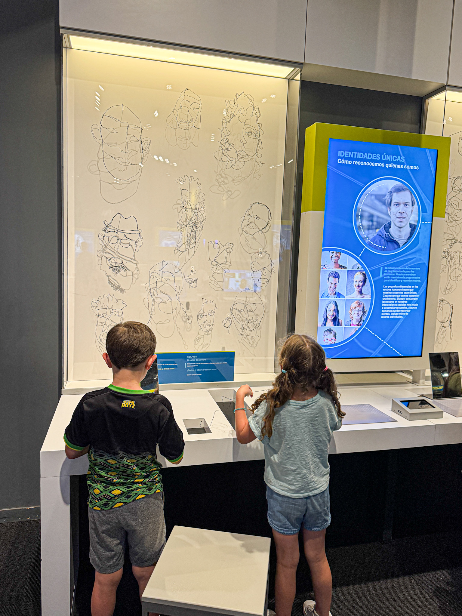 Two children observe an exhibit with abstract face sketches and an interactive display about unique identities.