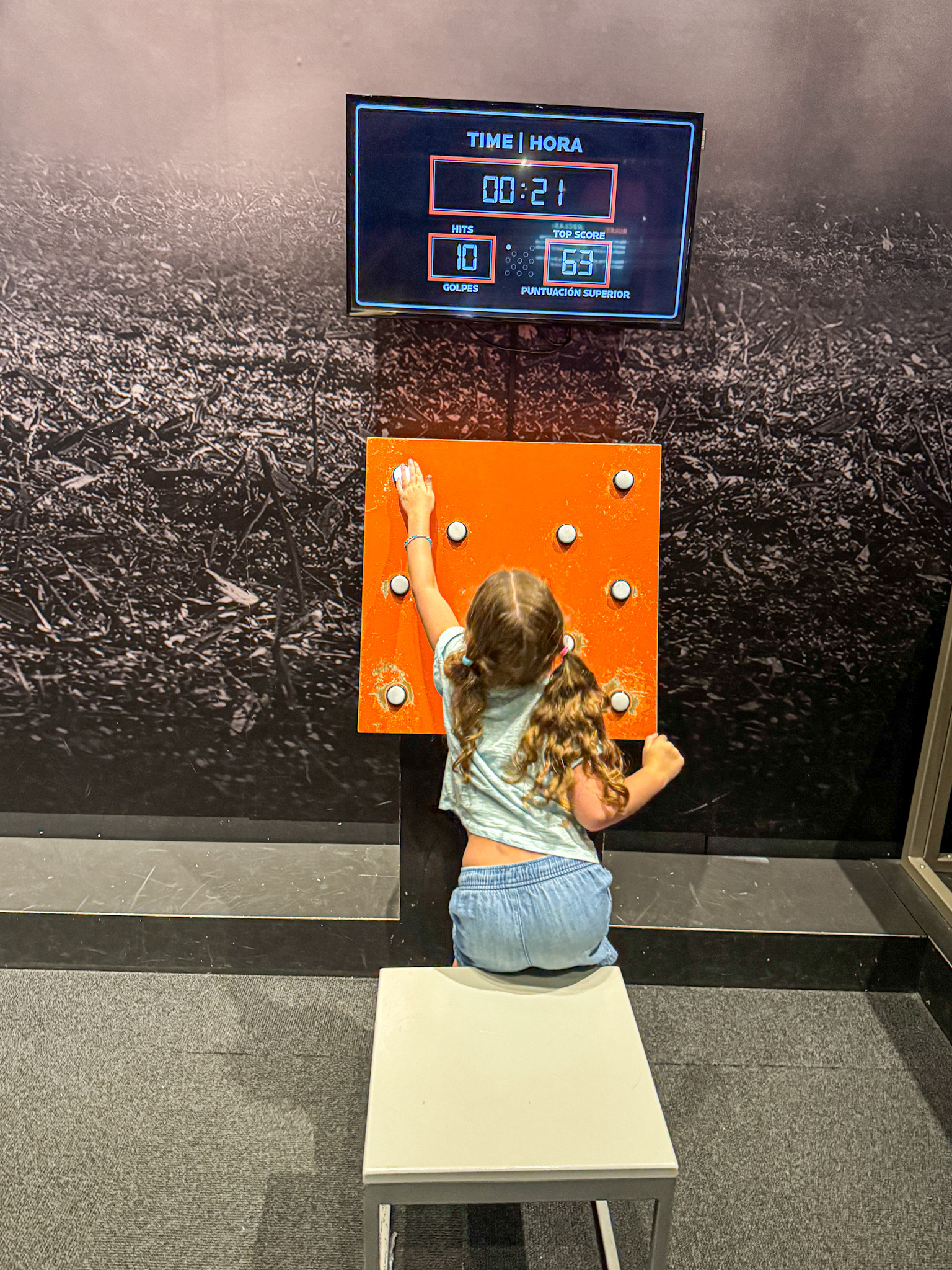 Young girl pressing buttons on an orange game panel beneath a digital scoreboard it the Sports Hall in a Dallas museum. 
