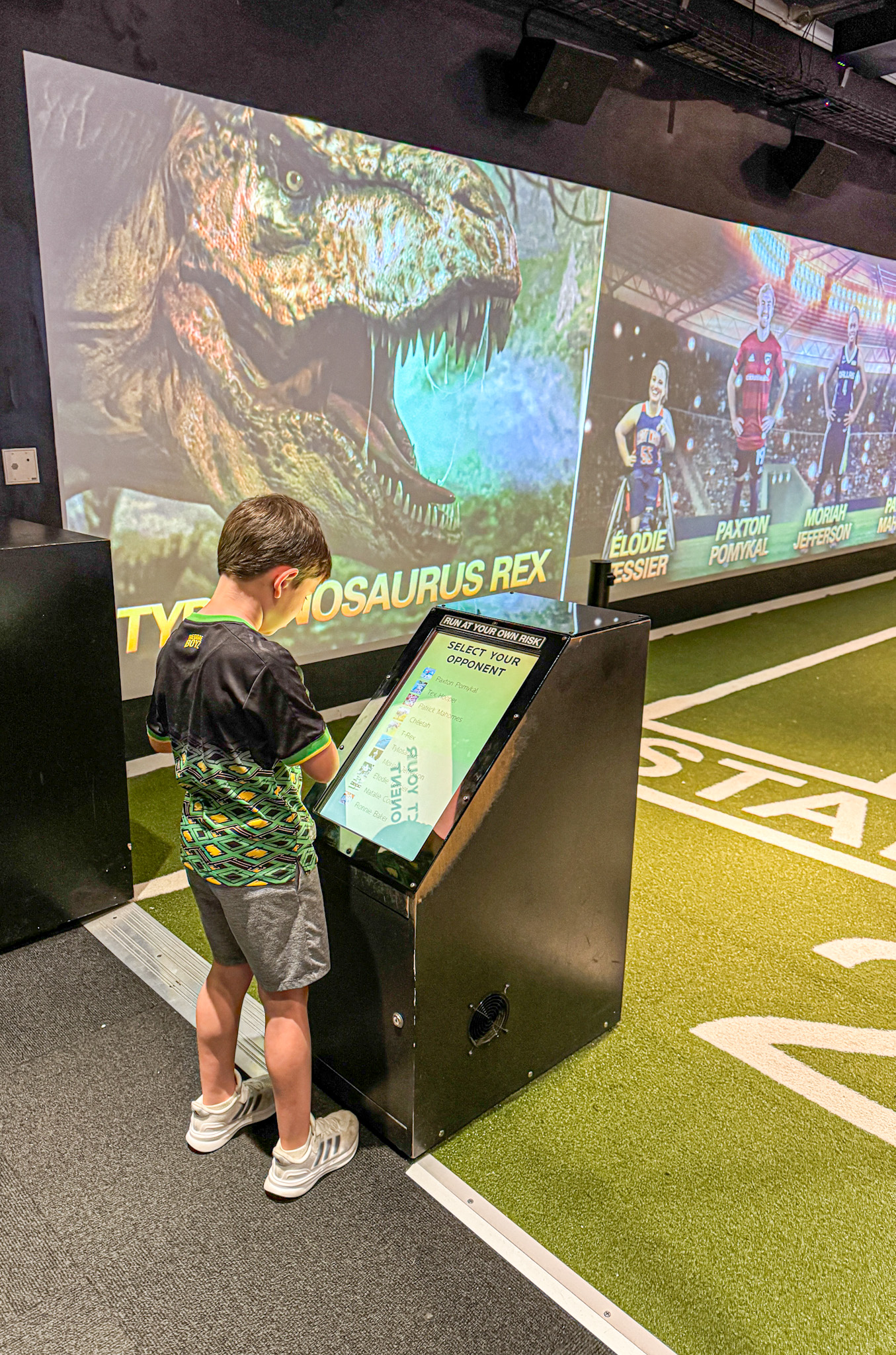 A young boy uses a touchscreen kiosk in a sports exhibit with dinosaur and athlete displays in the background.