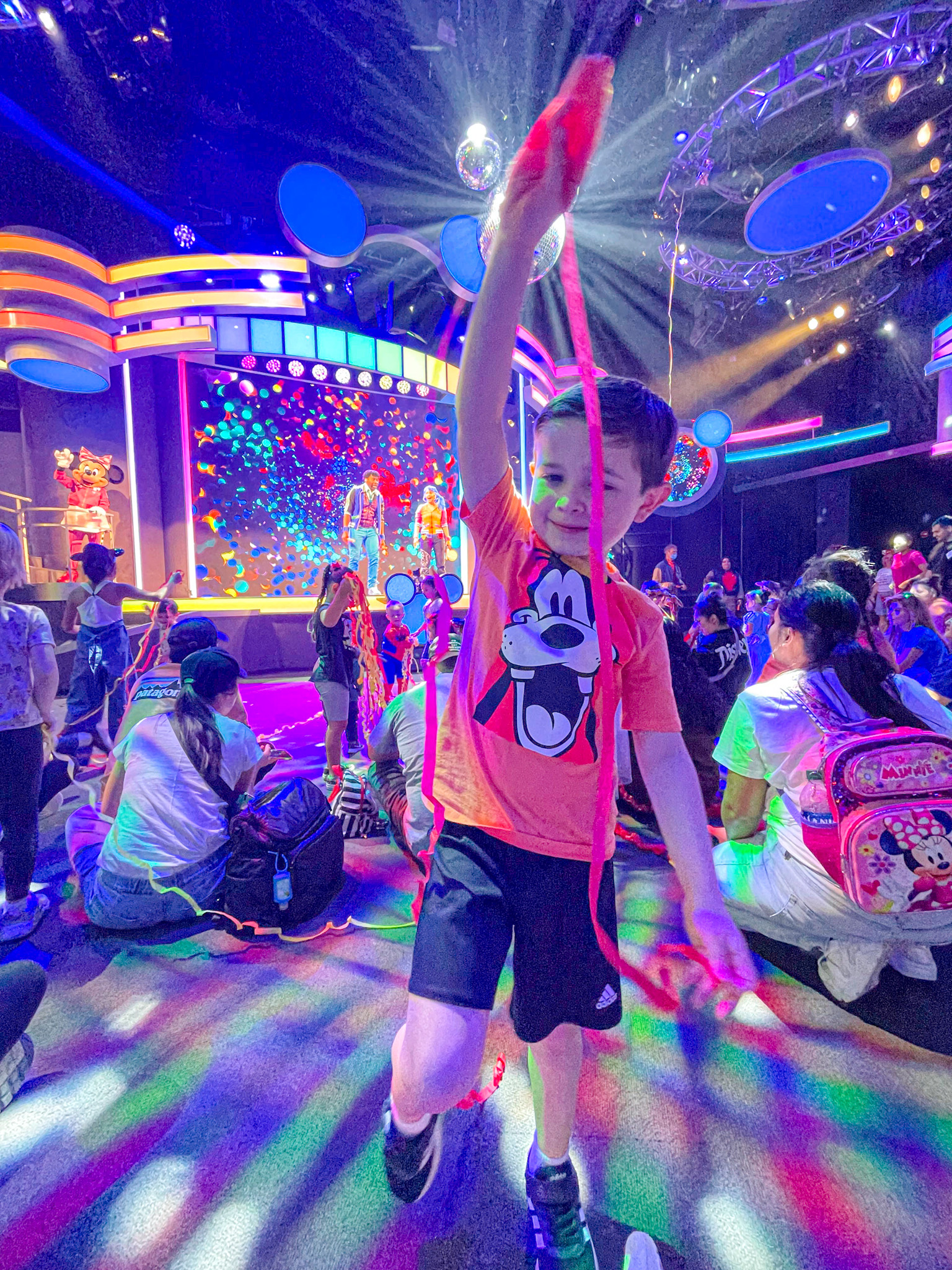 Young boy dances with streamers at a colorful indoor show, surrounded by seated audience and bright stage lights.