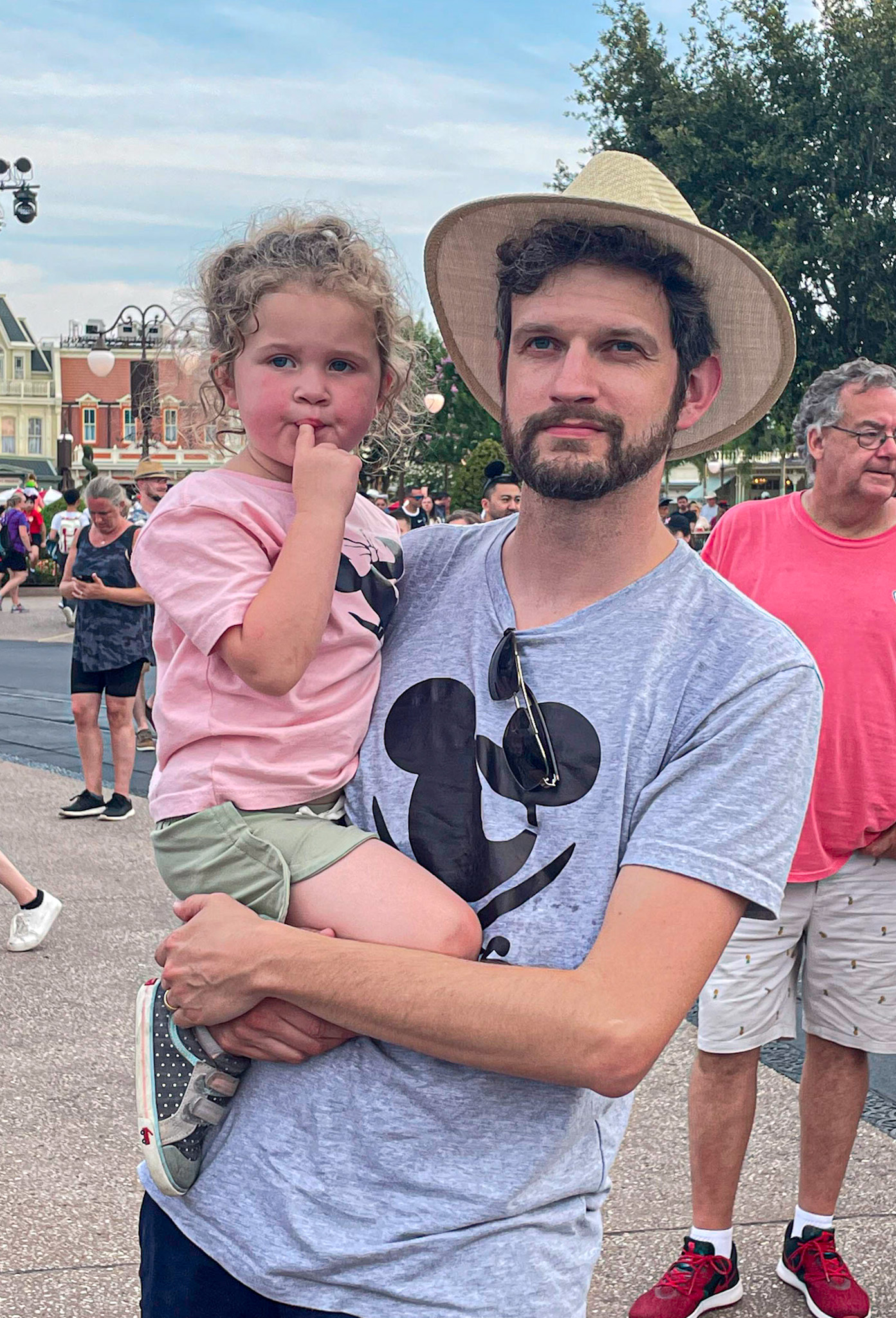 A man in a hat holds a young girl at a theme park, both wearing Mickey Mouse shirts.