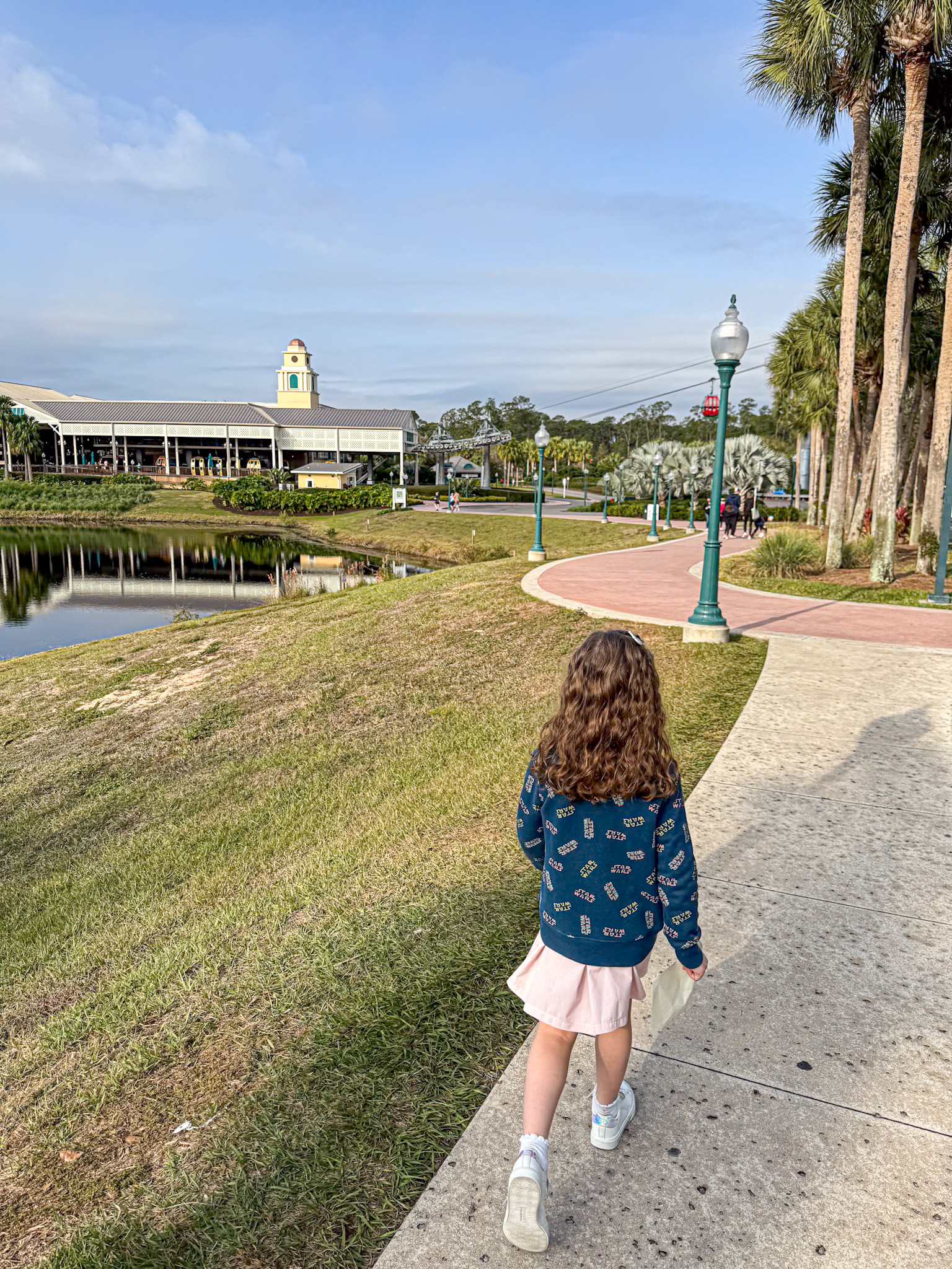 A young girl walks on a sidewalk by a pond, with trees and a building in the background on a sunny day.