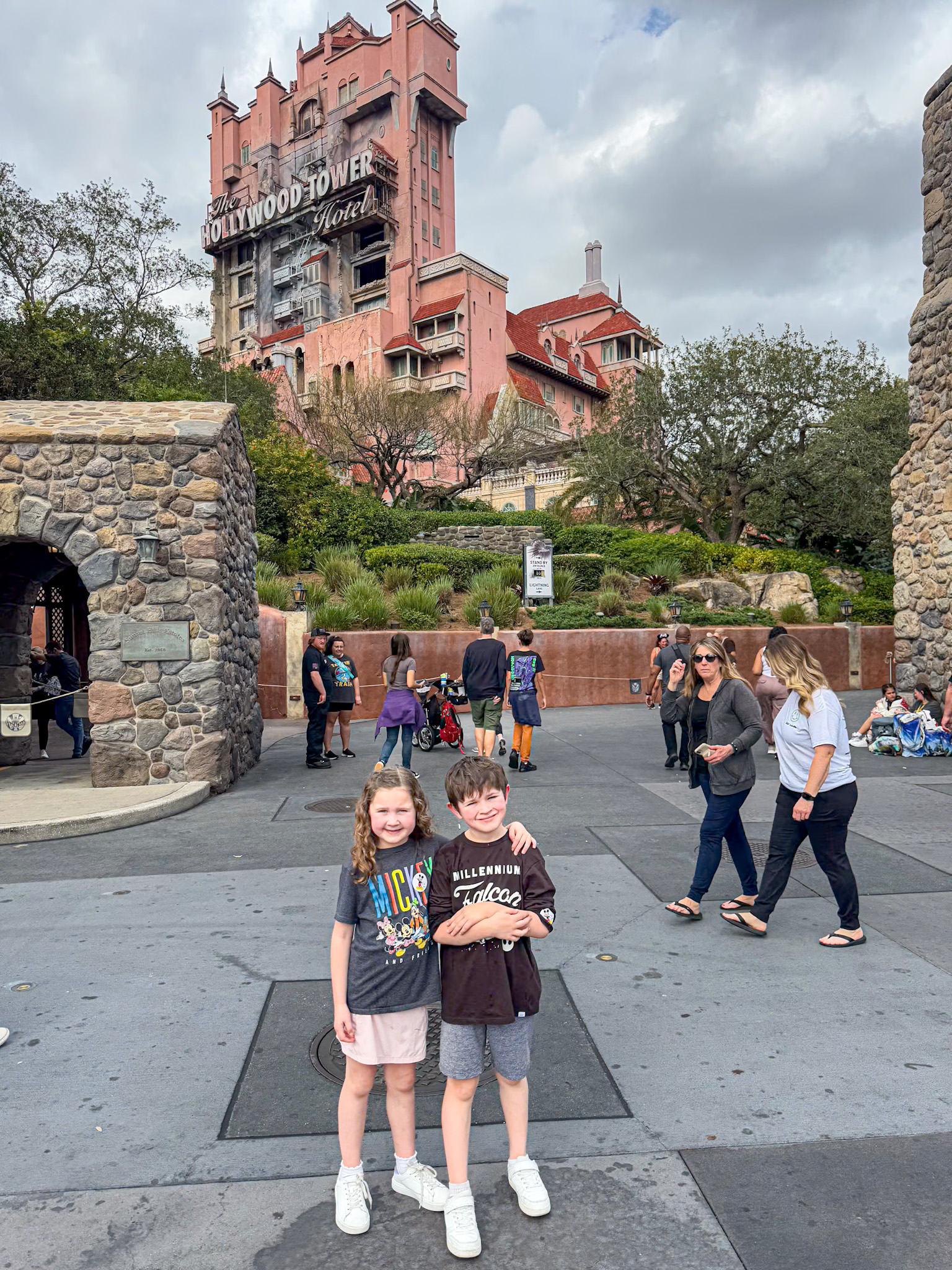 Two children smile in front of the Hollywood Tower Hotel at a Disney's Hollywood Studios theme park, with people walking nearby.