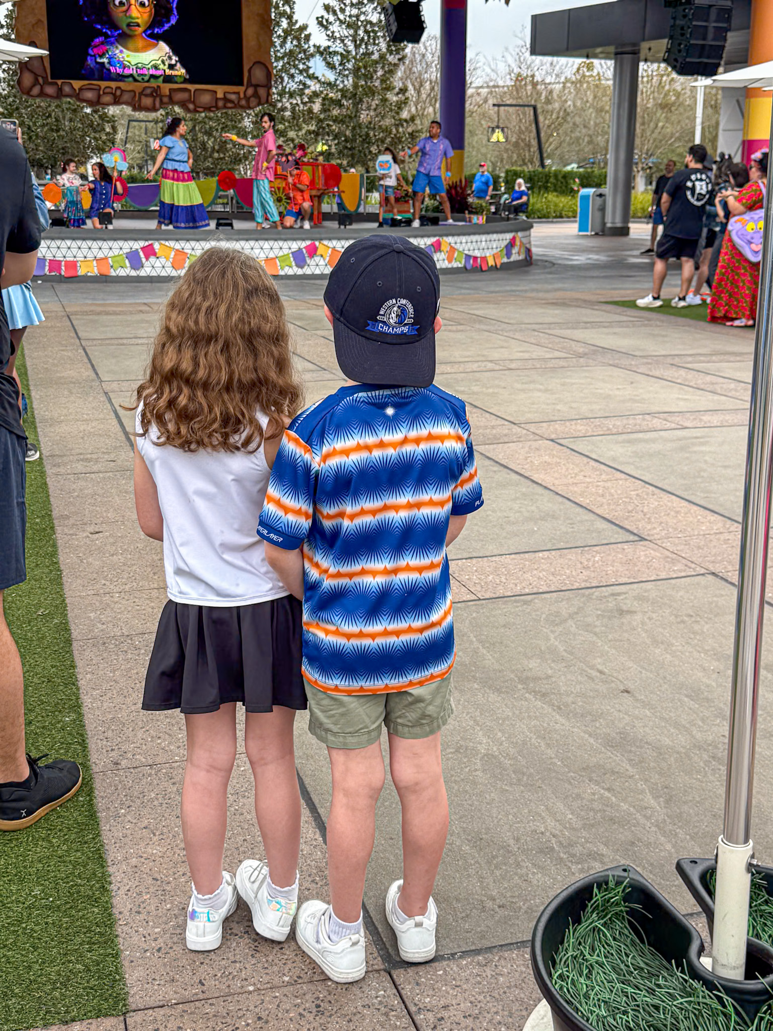 Two children stand side by side, watching a colorful outdoor stage performance with people and decorations.