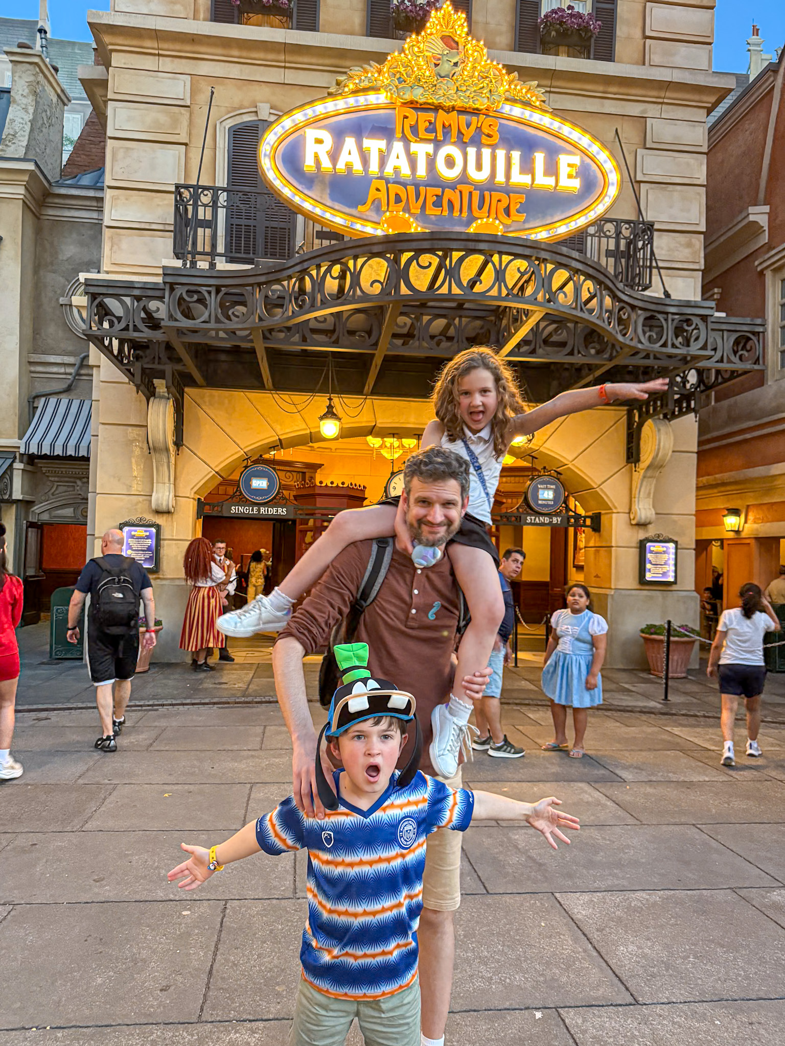 A father with two excited kids poses in front of Remys Ratatouille Adventure attraction at Epcot in Walt Disney World.