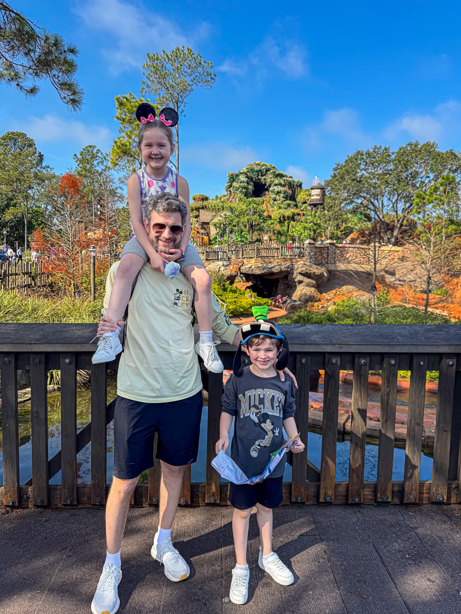 A man with two kids poses on a wooden bridge at Magic Kingdom with trees and a water ride in the background.