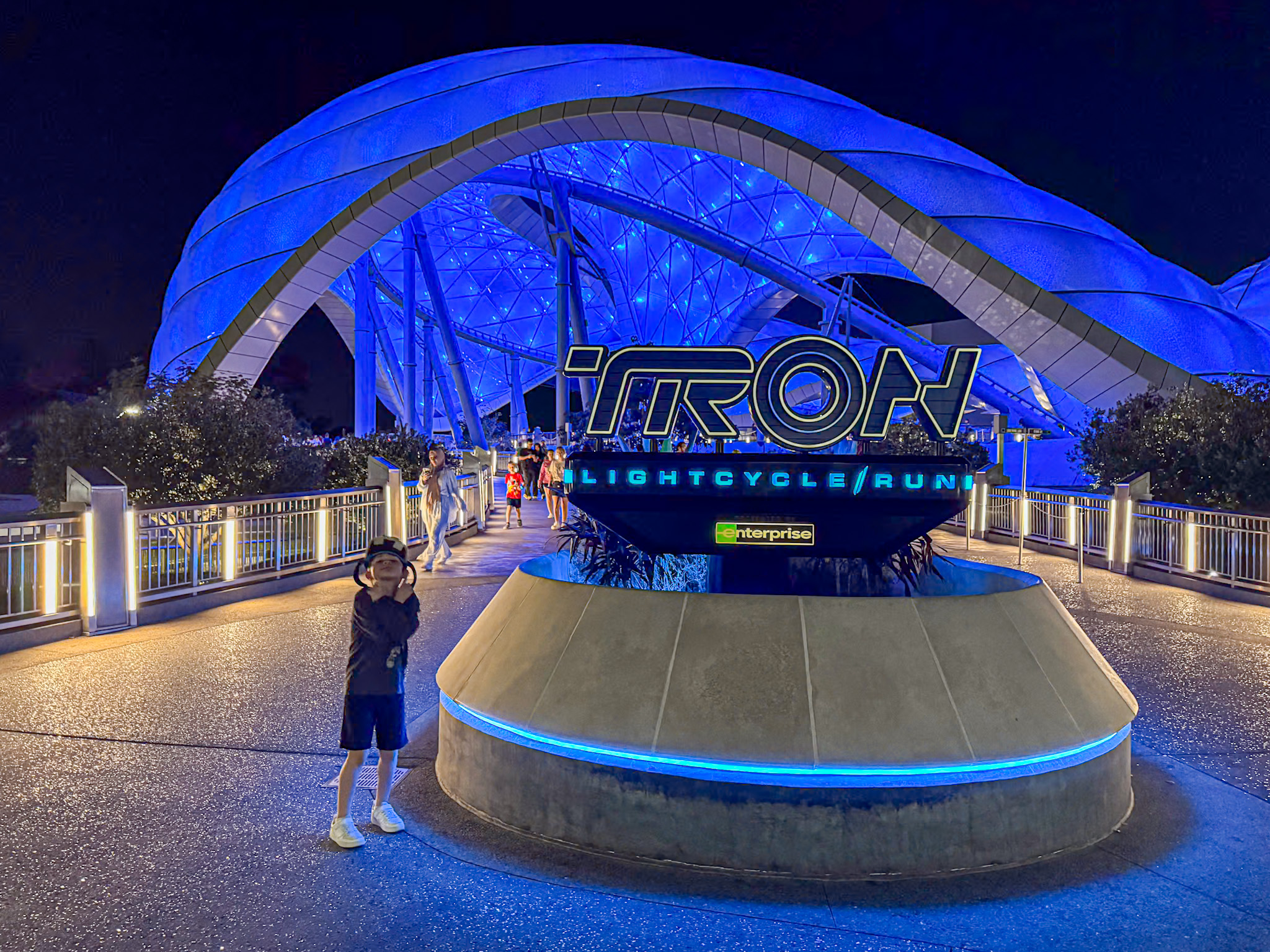 Child posing in front of the illuminated TRON Lightcycle / Run sign at night, under a blue-lit canopy at Disney World in Florida.