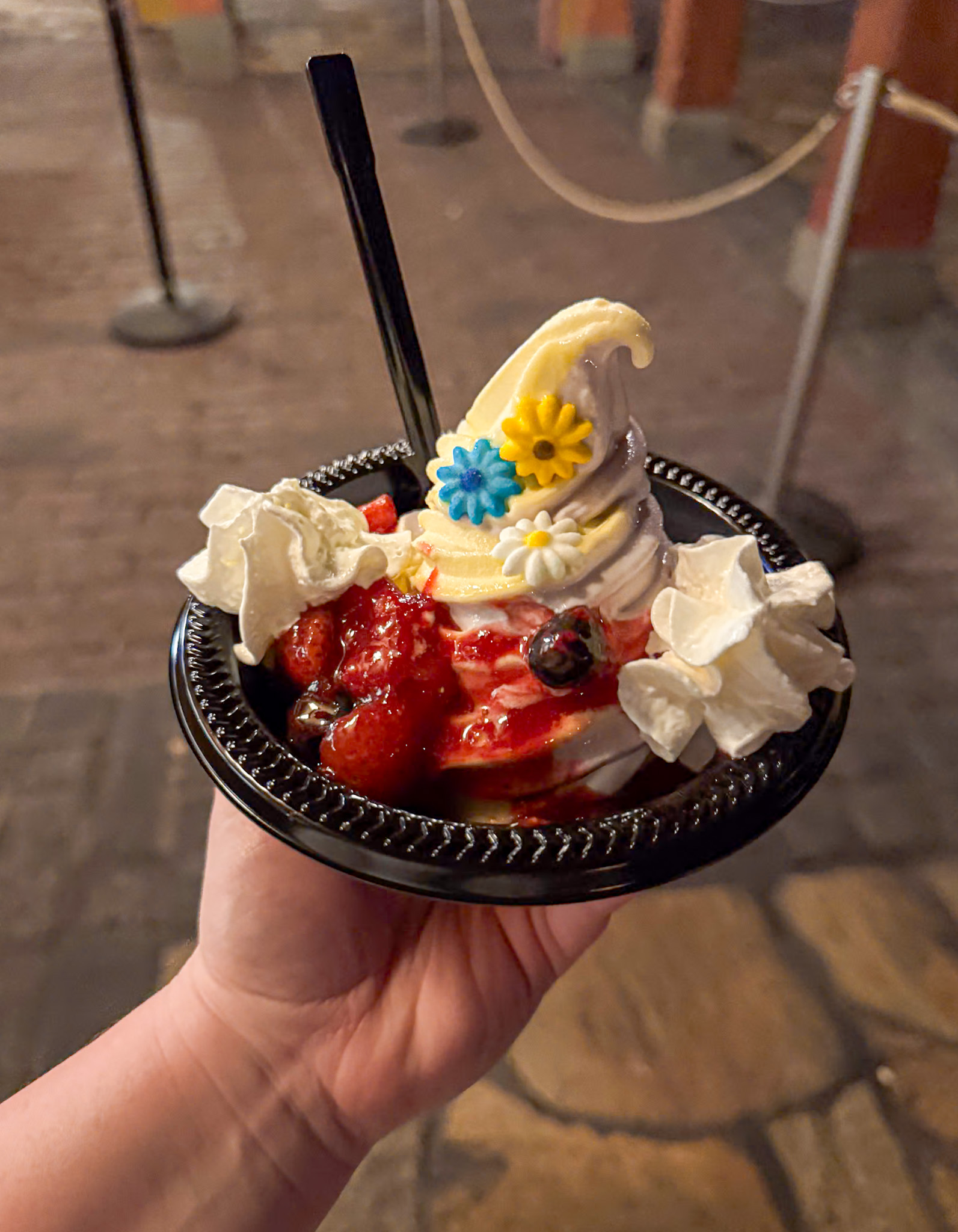Rapunzel soft serve ice cream sundae with berry sauce, whipped cream, and flower decorations in a black bowl, held in hand.