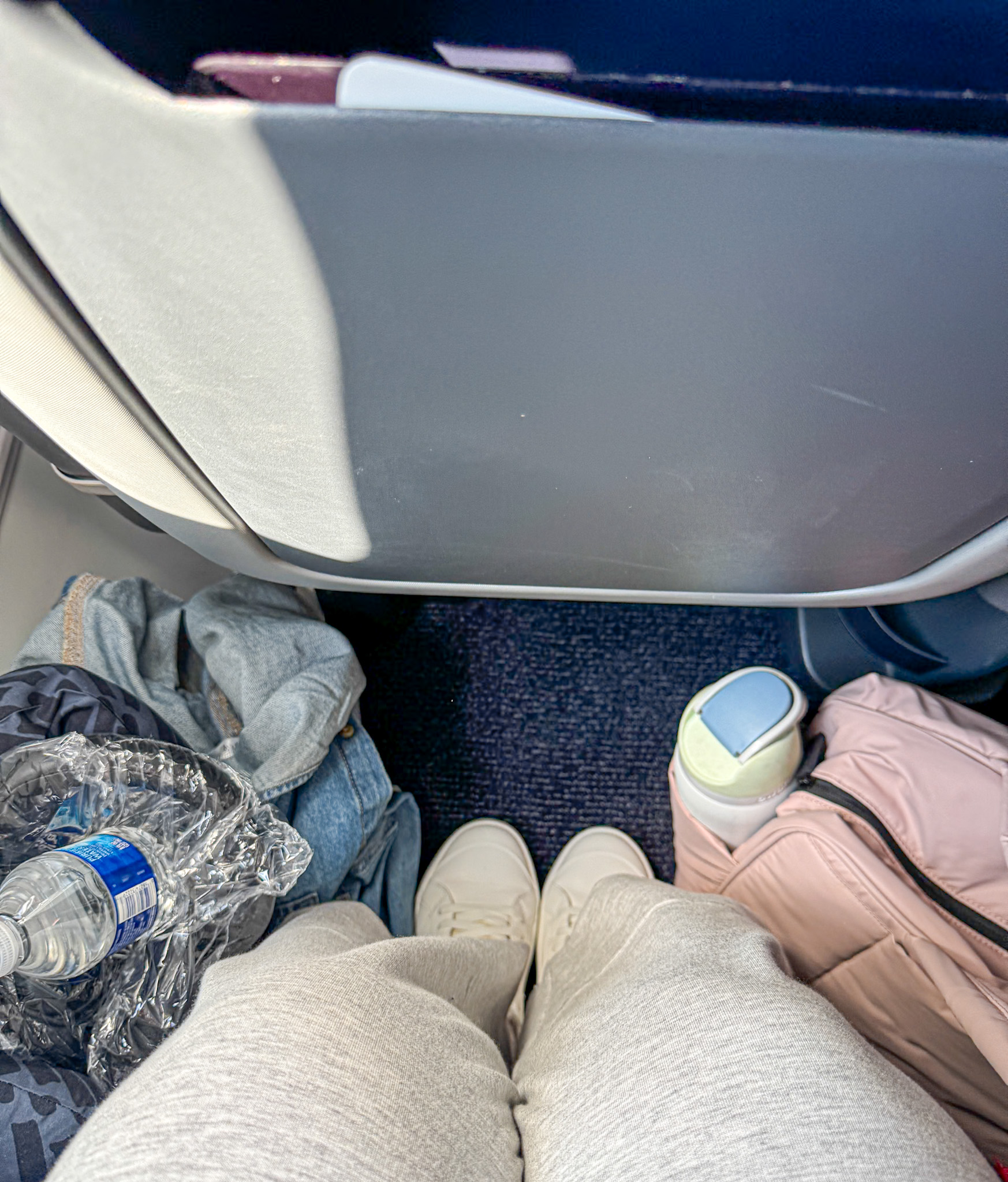 Legs in sweatpants with a backpack, water bottle, and crumpled plastic in an airplane seats legroom area.