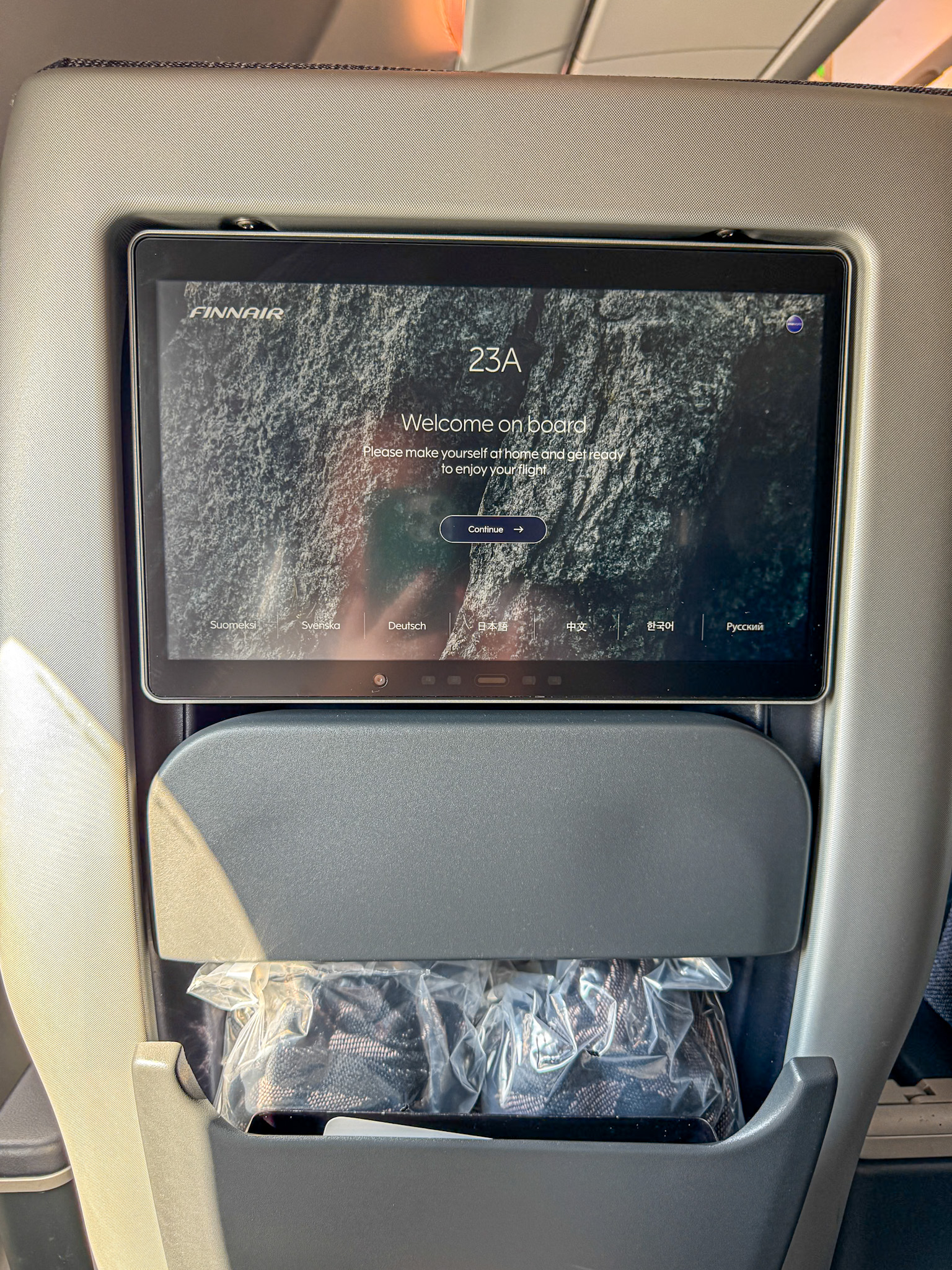 Finnair Premium Economy airplane seatback screen showing a welcome message, with headphones in a plastic bag in the pocket below.