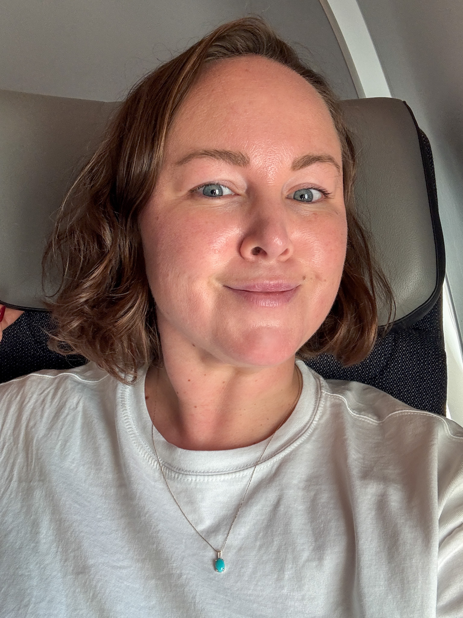 Woman with short brown hair wearing a white shirt and turquoise necklace, smiling on a Finnair airplane seat.