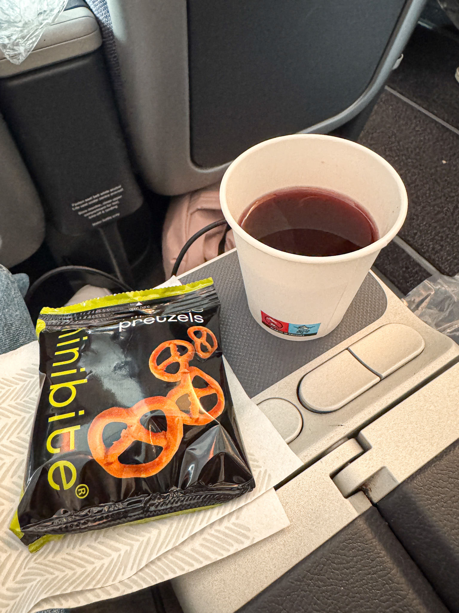 A cup of blueberry juice and a bag of pretzels on an airplane tray table.