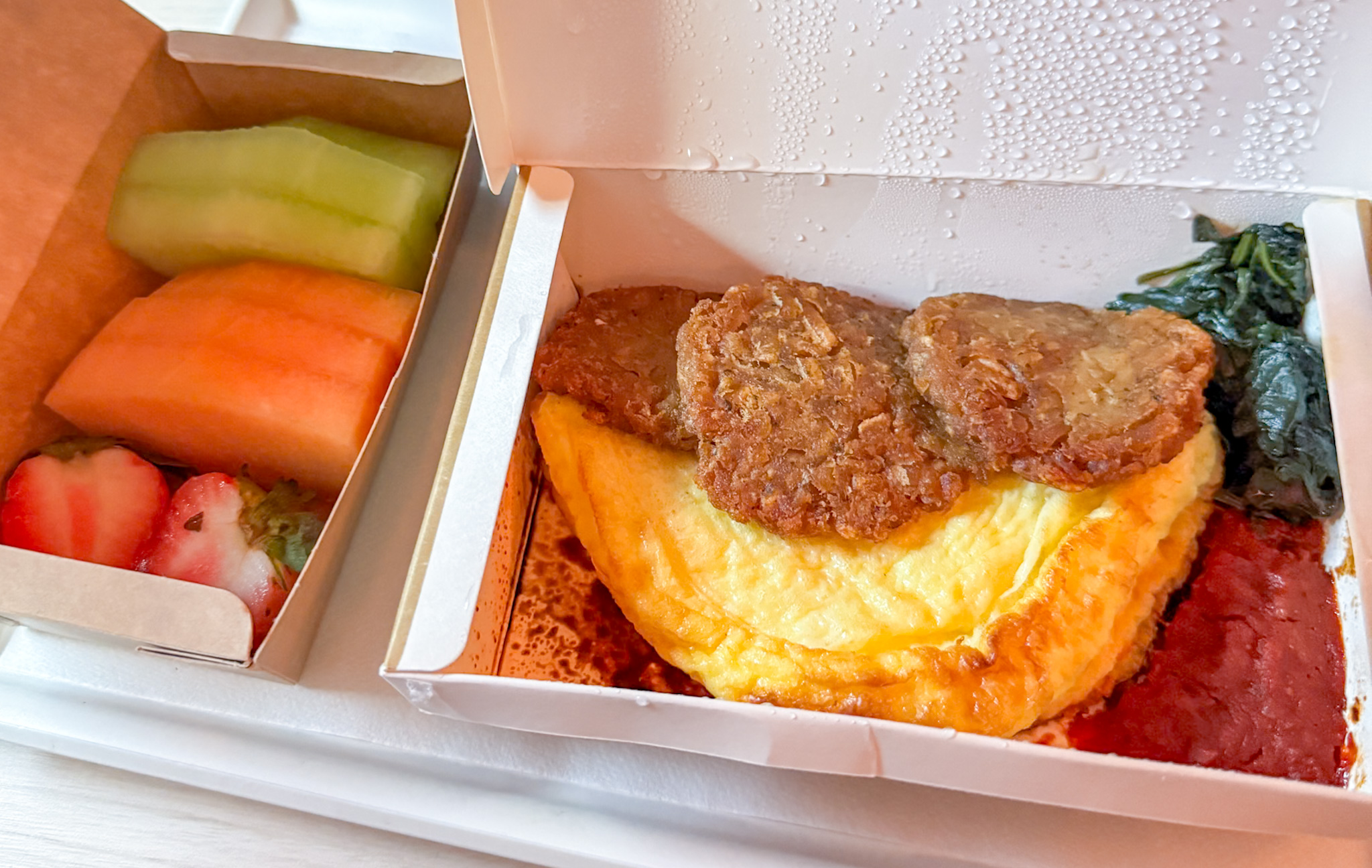 Boxed airline meal with omelette, sausage patties, spinach, tomato sauce, and a side of sliced fruit and strawberries.