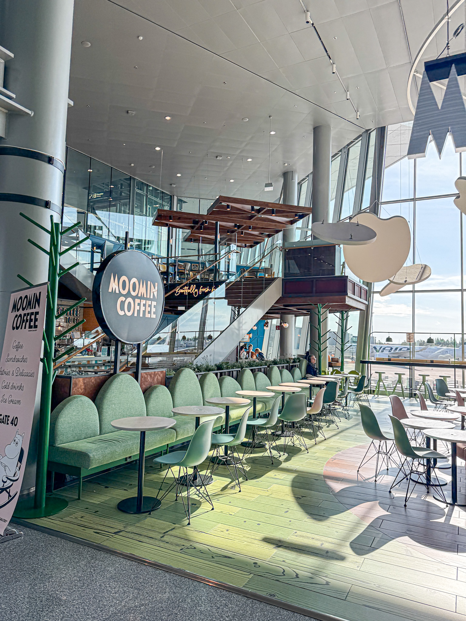 Bright, modern café with green seating, Moomin Coffee sign, large windows, and whimsical Moomin decor at Helsinki airport in Finland.
