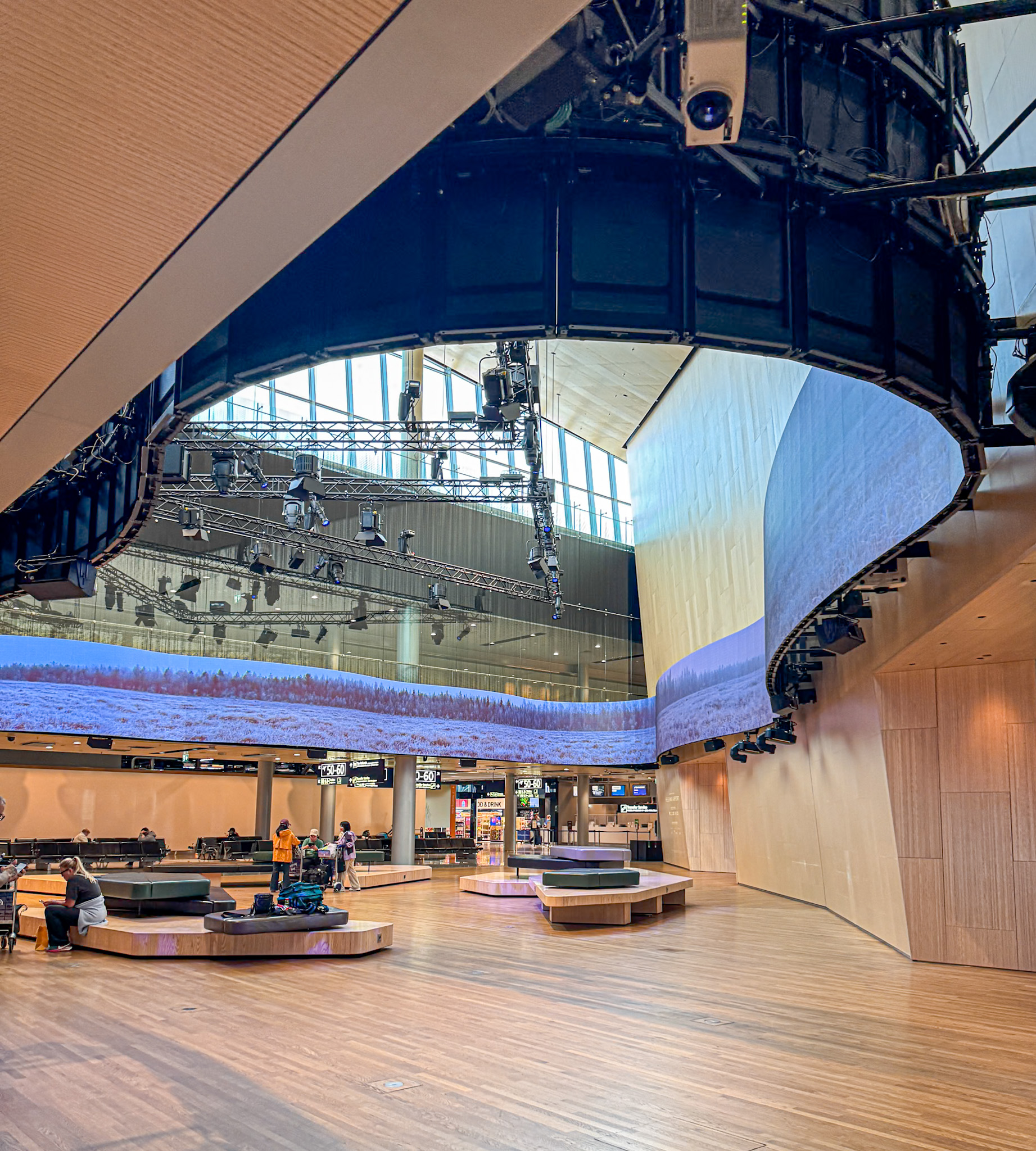 Spacious modern lobby in Helsinki airport with curved wooden walls, large screen, seating areas, and high windows.