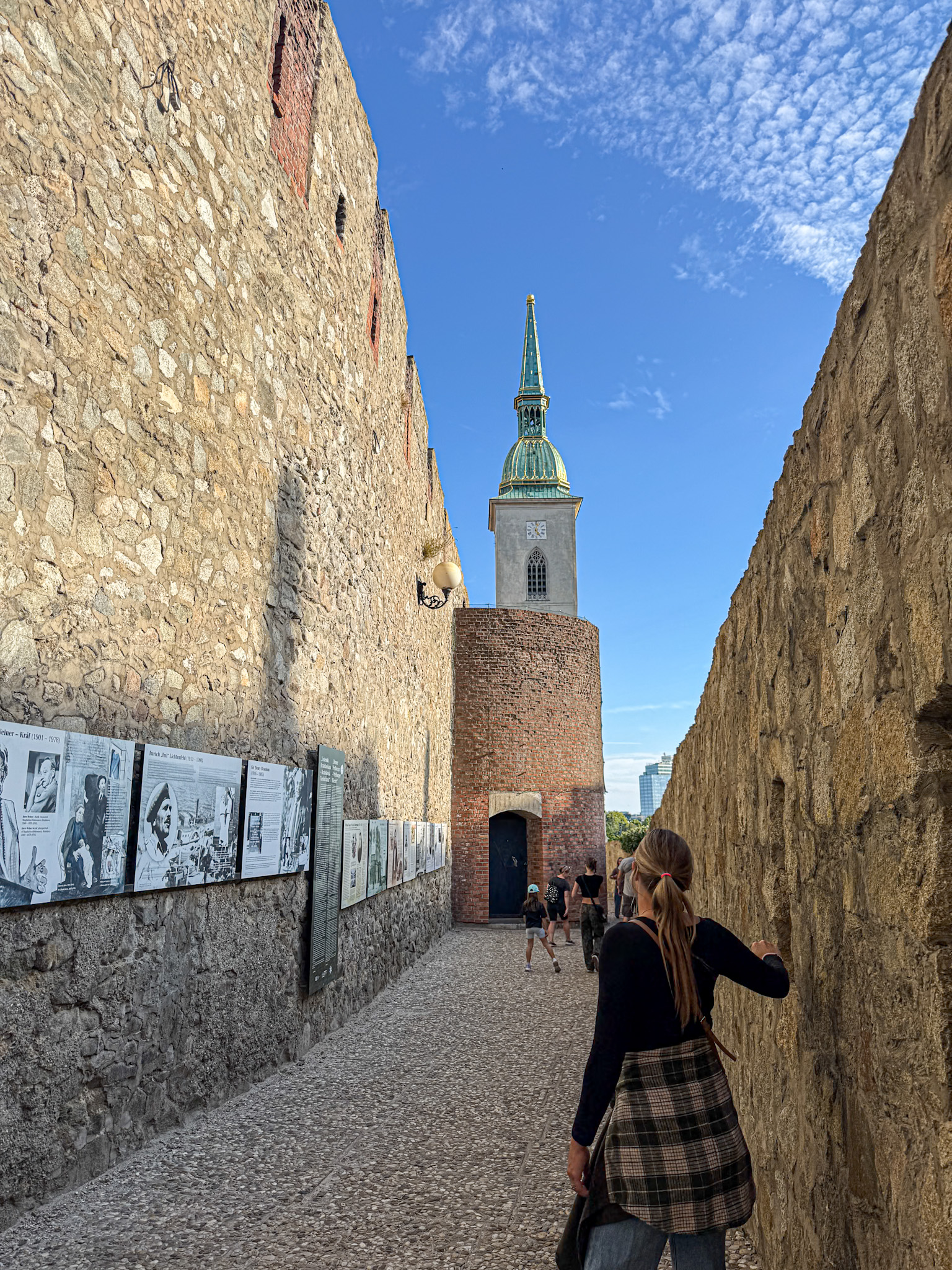 A woman walks down a narrow stone alley towards a church tower under a blue sky with scattered clouds.